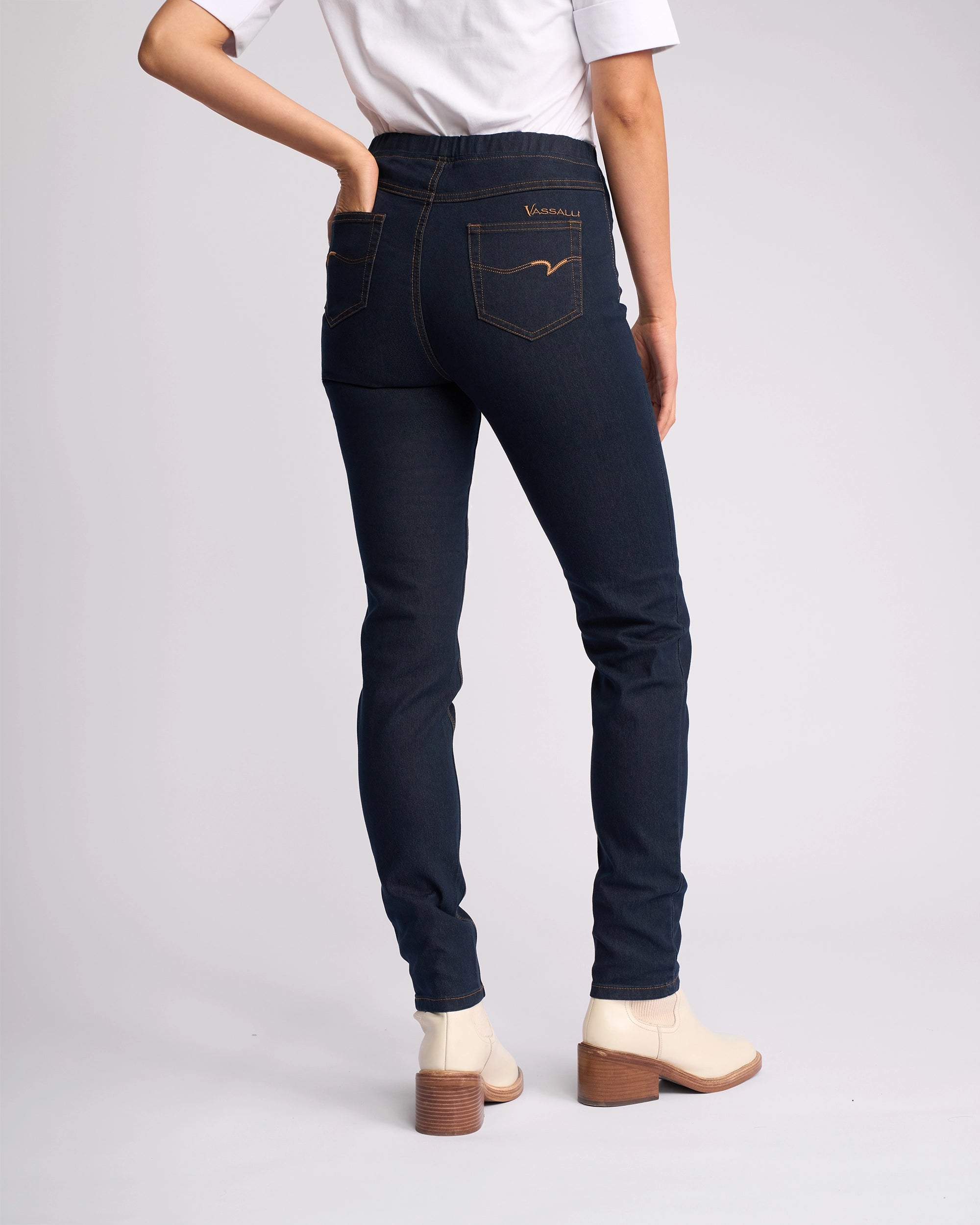 Wearing Vassalli's Origin Pull On - Indigo Denim (Contrast Stitch) jeans, paired with a white top and light ankle boots with a brown heel, a person stands facing away from the camera with one hand in their back pocket.