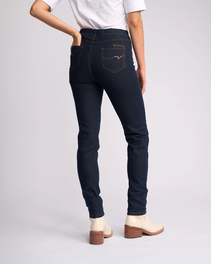 Wearing Vassalli's Origin Pull On - Indigo Denim (Contrast Stitch) jeans, paired with a white top and light ankle boots with a brown heel, a person stands facing away from the camera with one hand in their back pocket.