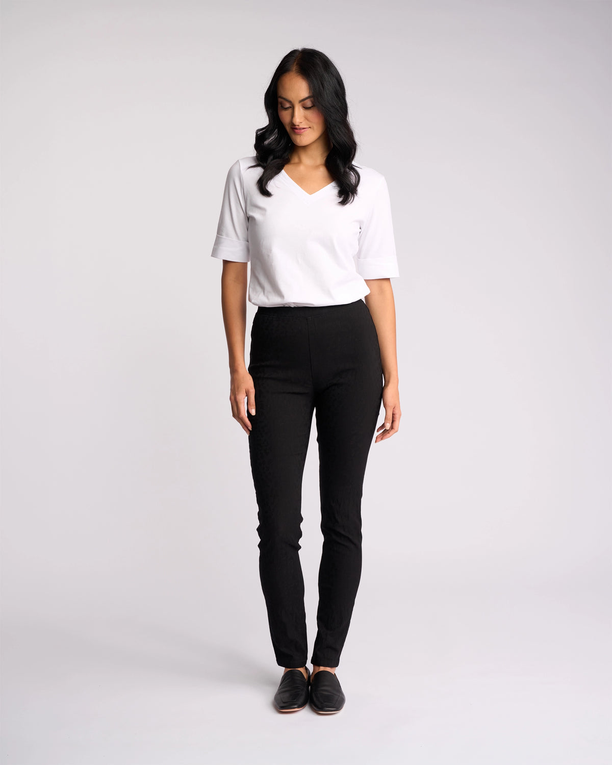 A woman with long dark hair, wearing a white short-sleeve V-neck shirt and Vassalli Origin Pull On - Black Embossed pants, stands against a plain white background, looking down with a slight smile.