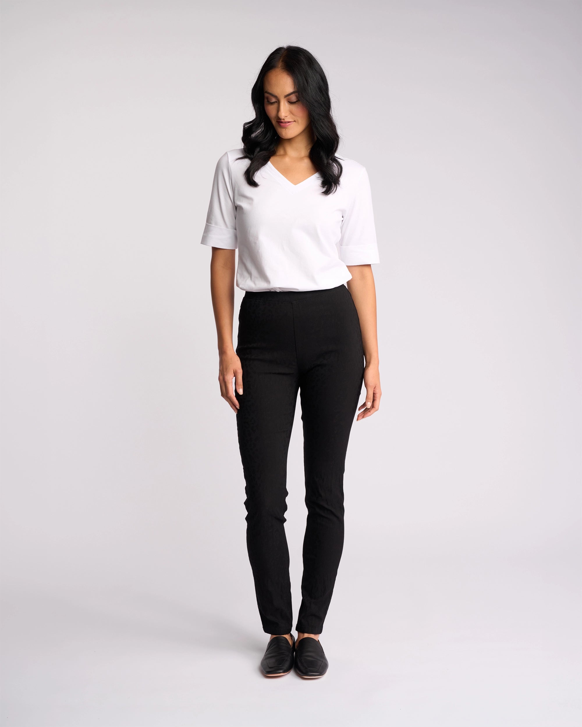 A woman with long dark hair, wearing a white short-sleeve V-neck shirt and Vassalli Origin Pull On - Black Embossed pants, stands against a plain white background, looking down with a slight smile.
