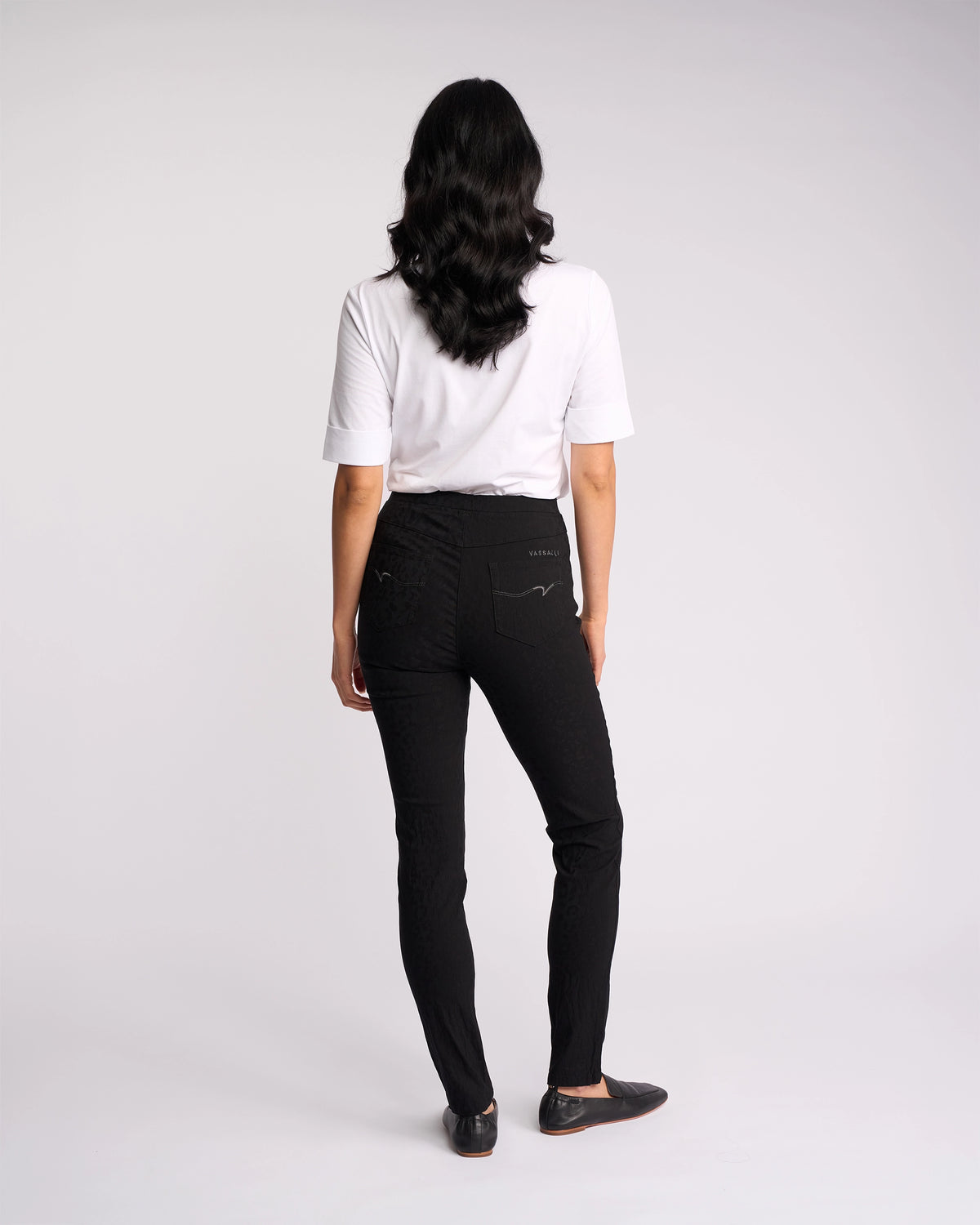 A person with long dark hair, wearing a white short-sleeve shirt and Vassalli Origin Pull On - Black Embossed pants, stands facing away from the camera against a plain light background.