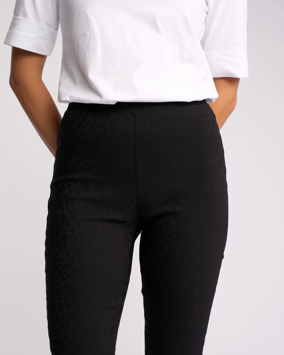 A person stands with hands behind their back, shown from shoulders to knees against a plain background, wearing a white short-sleeve shirt and Vassalli's Origin Pull On - Black Embossed pants.