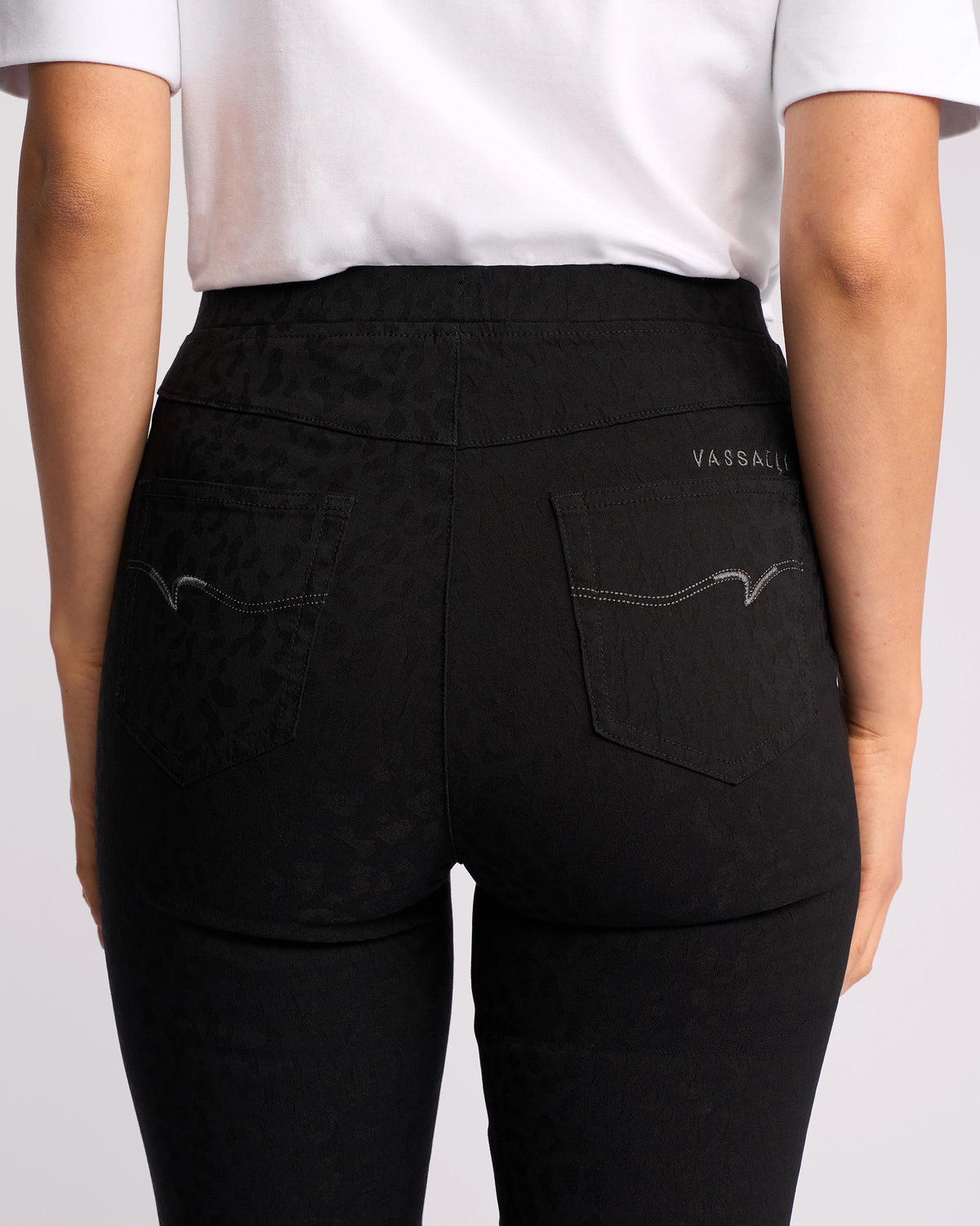 A person wearing a white shirt and Vassalli Origin Pull On - Black Embossed pants, viewed from the back. The high-waisted skinny jeans feature two back pockets with decorative stitching and VASSALLI embroidered on one pocket.
