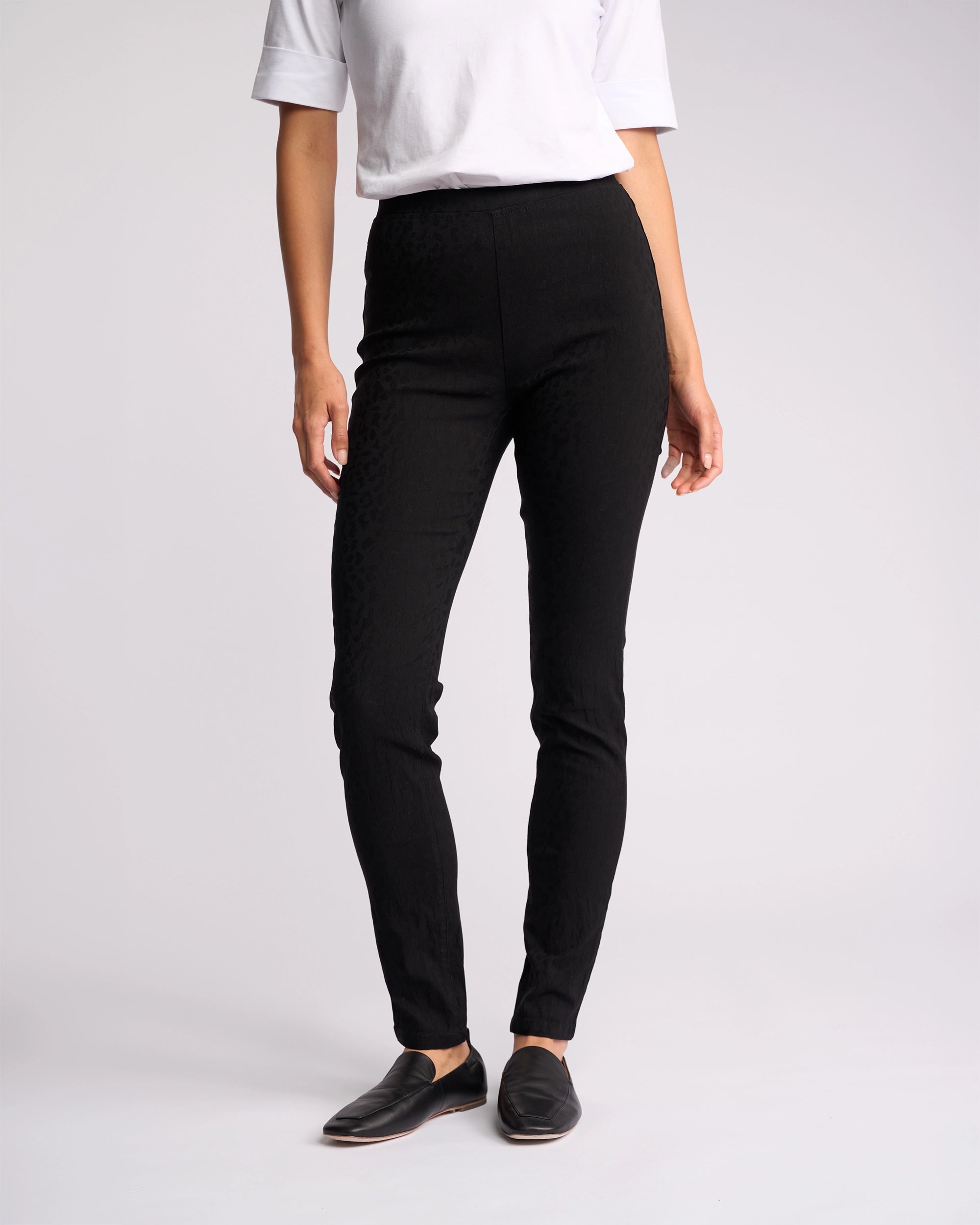 A person stands against a plain light background, wearing a white short-sleeve top, Vassalli Origin Pull On - Black Embossed high rise skinny-leg pants, and black slip-on shoes. Their head is not visible in the image.