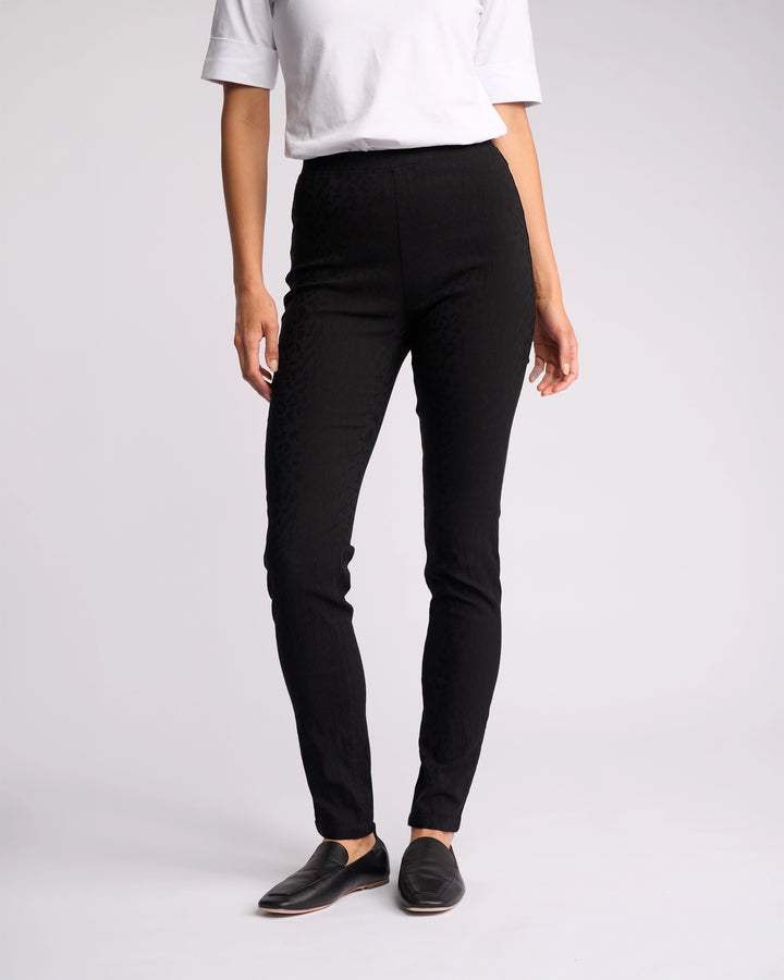 A person stands against a plain light background, wearing a white short-sleeve top, Vassalli Origin Pull On - Black Embossed high rise skinny-leg pants, and black slip-on shoes. Their head is not visible in the image.