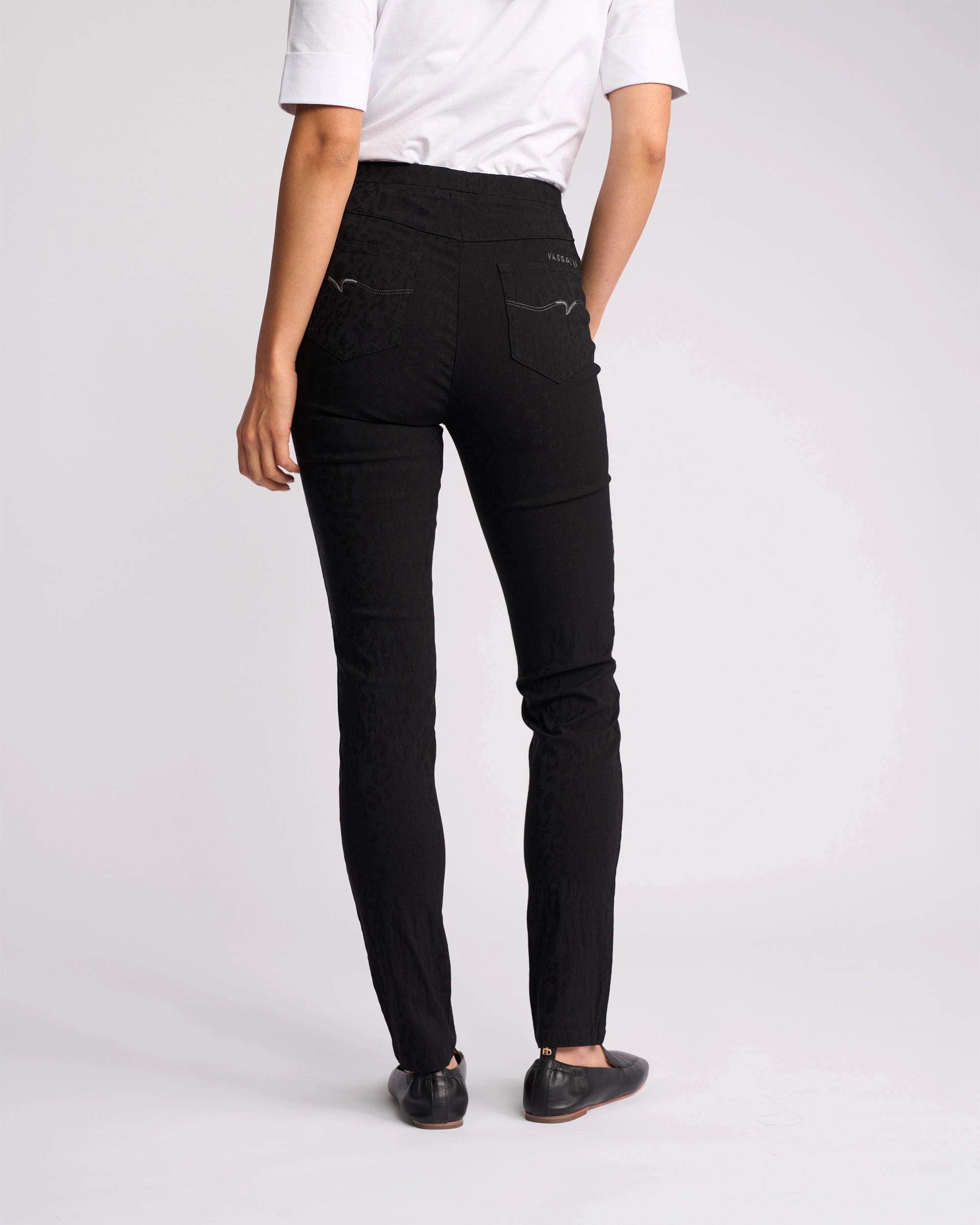 A person faces away, wearing a white short-sleeve shirt and Vassalli Origin Pull On - Black Embossed skinny-leg pants, paired with black flats, against a plain light background.