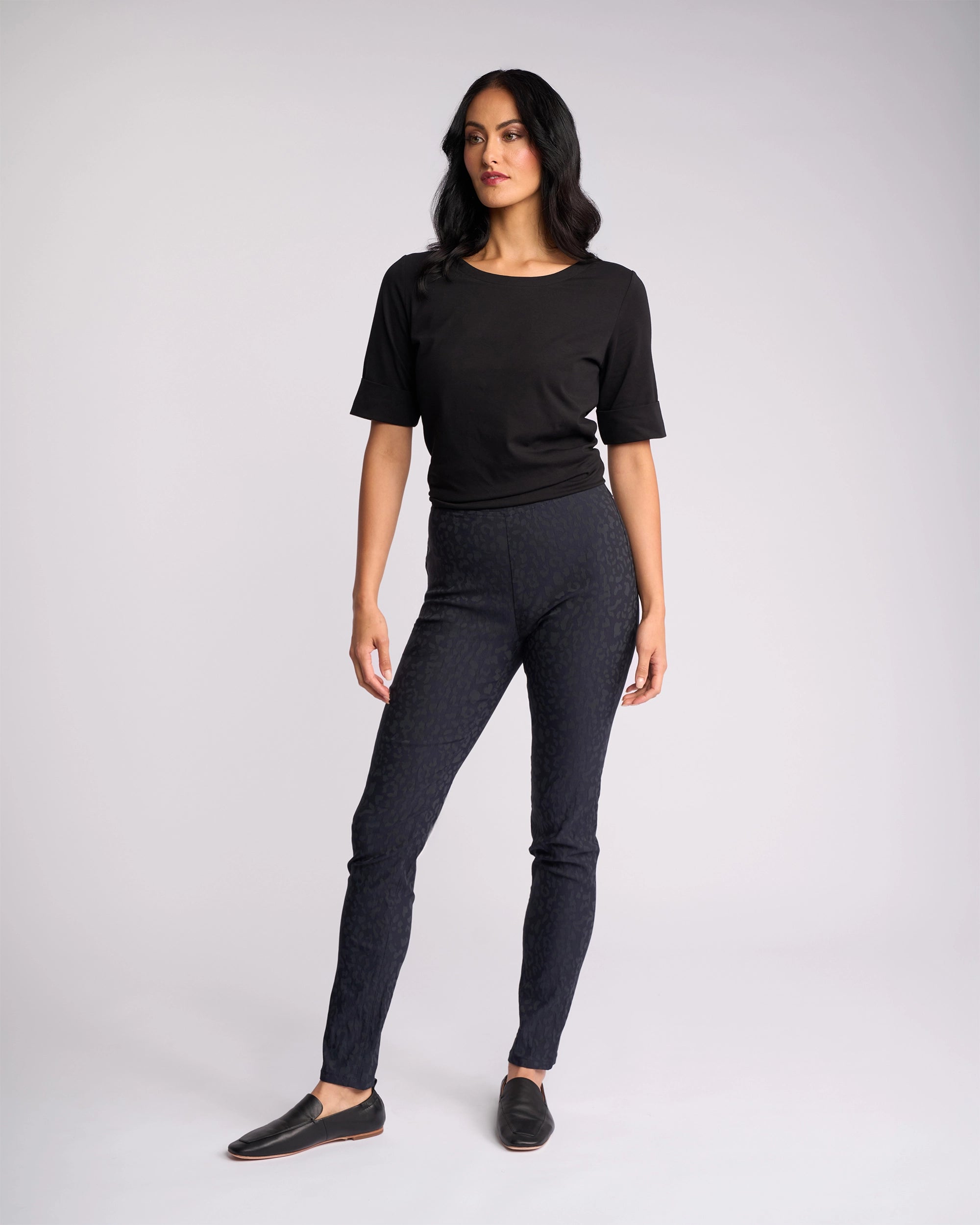 A woman with long dark hair stands against a plain background wearing Vassalli's Origin Pull On - Navy Embossed high rise skinny-leg pants, a black short-sleeve top, and black slip-on shoes, looking slightly to the side with a relaxed posture.
