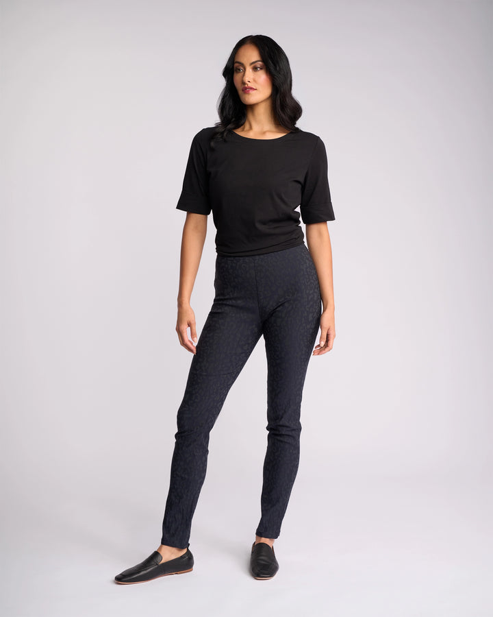 A woman with long dark hair stands against a plain background wearing Vassalli's Origin Pull On - Navy Embossed high rise skinny-leg pants, a black short-sleeve top, and black slip-on shoes, looking slightly to the side with a relaxed posture.