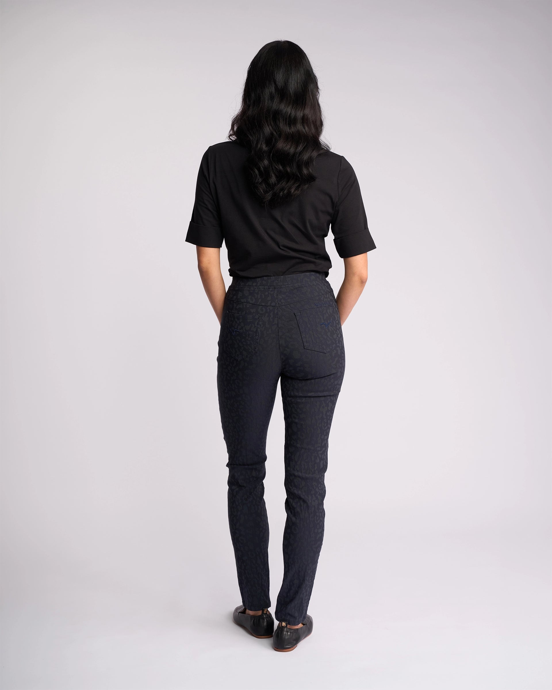 A person with long dark hair faces away wearing Vassalli’s Origin Pull On - Navy Embossed pants and a black short-sleeve shirt with black shoes, standing against a light gray background.