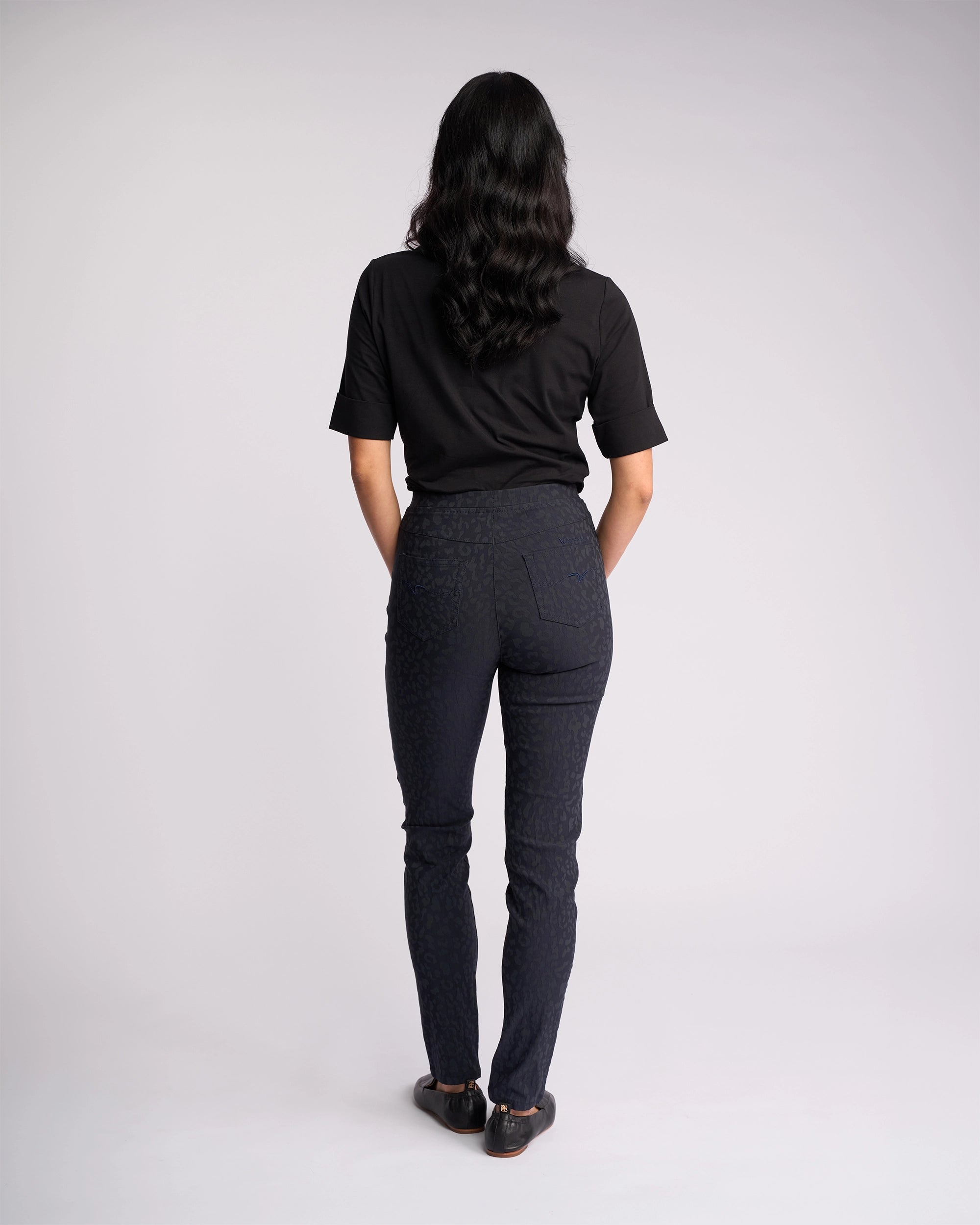 A person with long dark hair faces away wearing Vassalli’s Origin Pull On - Navy Embossed pants and a black short-sleeve shirt with black shoes, standing against a light gray background.