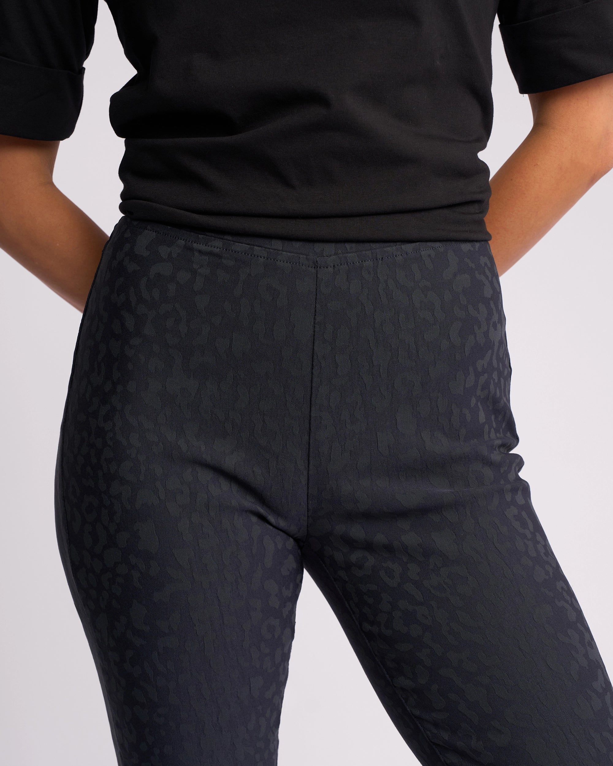 A person in a black short-sleeved top and Vassalli Origin Pull On - Navy Embossed high-rise, skinny-leg pants stands with arms behind their back against a plain light background.