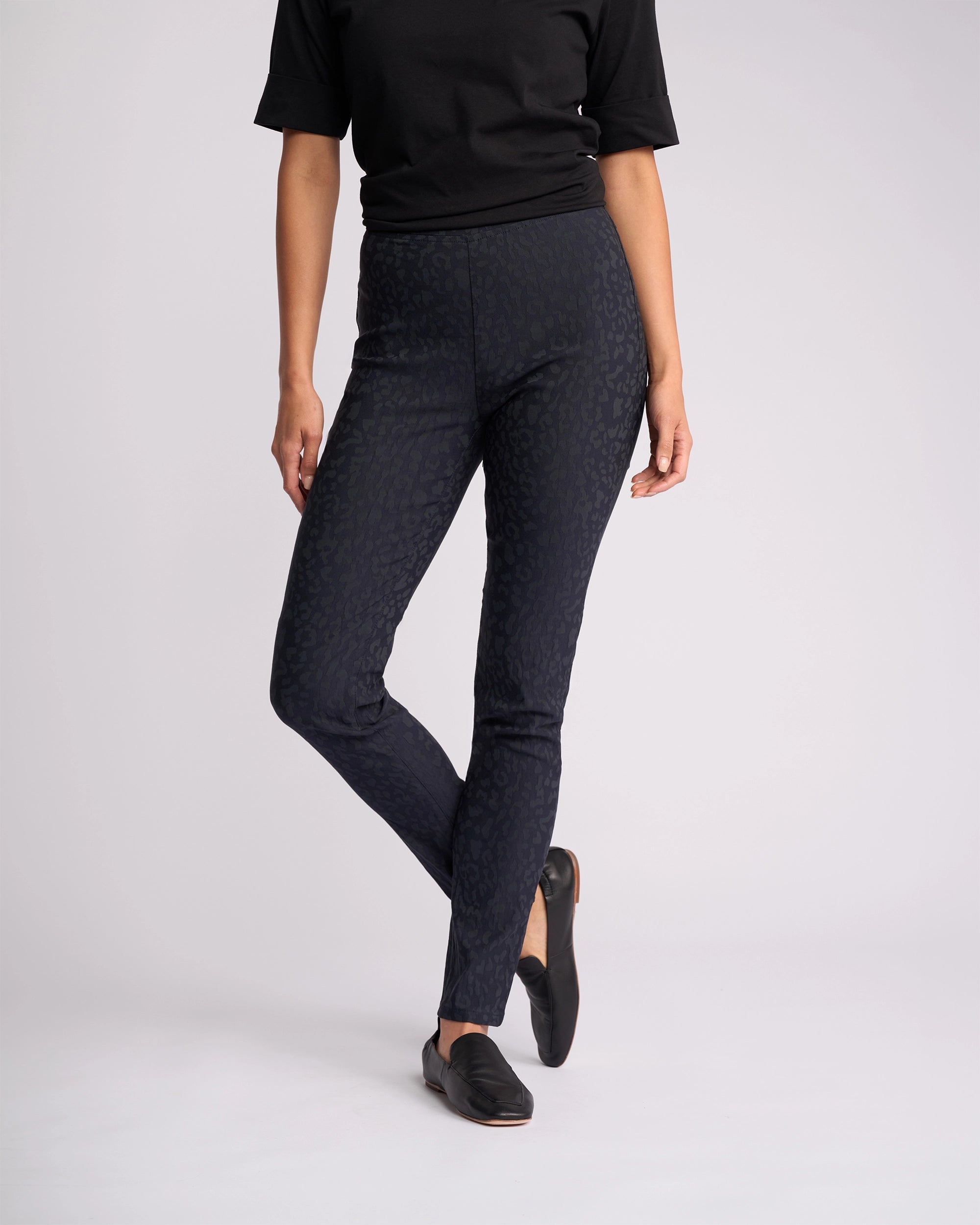A person stands against a plain light background, one leg slightly crossed, wearing a black short-sleeve top, black loafers, and Vassalli Origin Pull On - Navy Embossed skinny-leg pants.