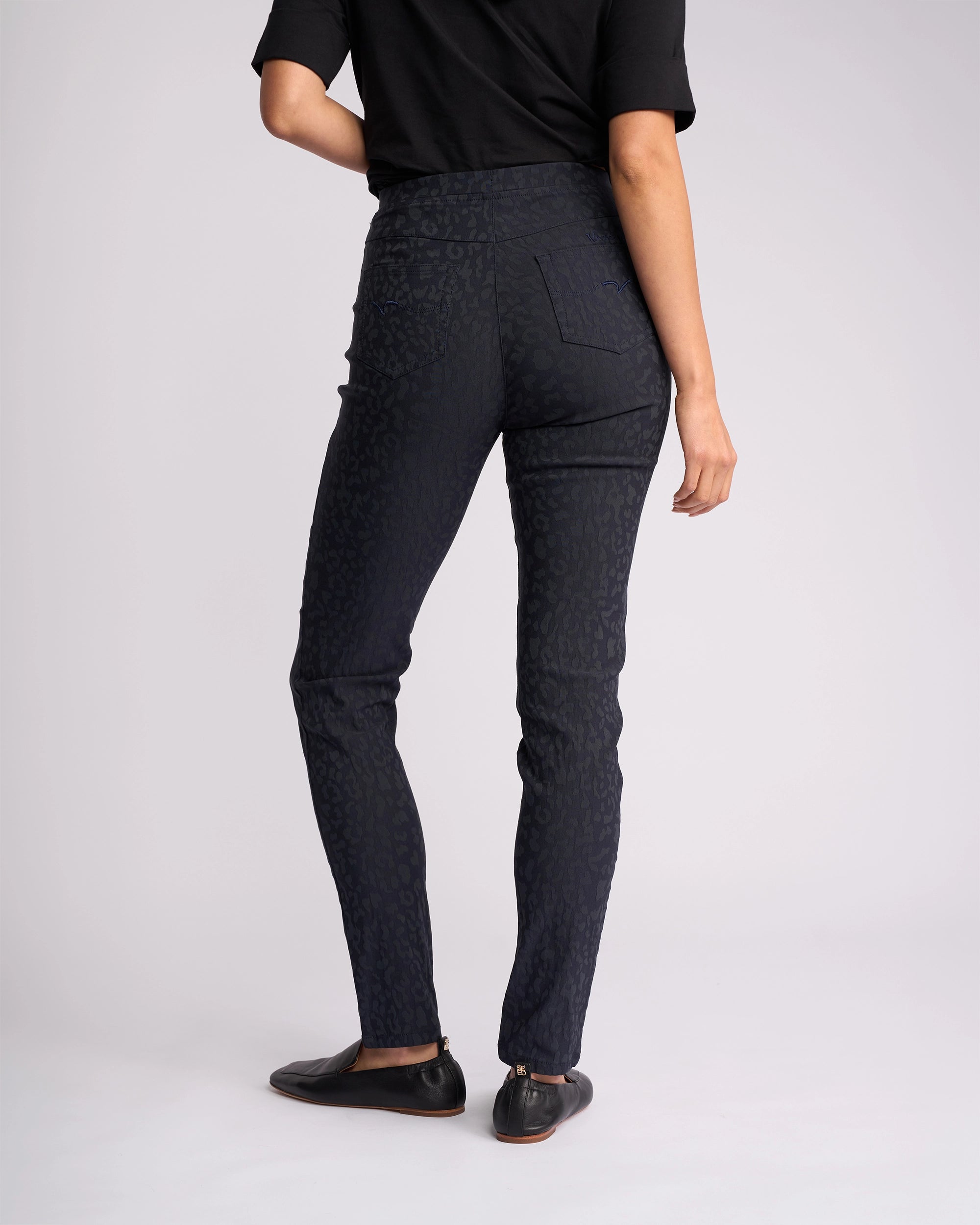 A person stands facing away, wearing a black short-sleeve top and Vassalli Origin Pull On - Navy Embossed pants, paired with black slip-on shoes against a light background.