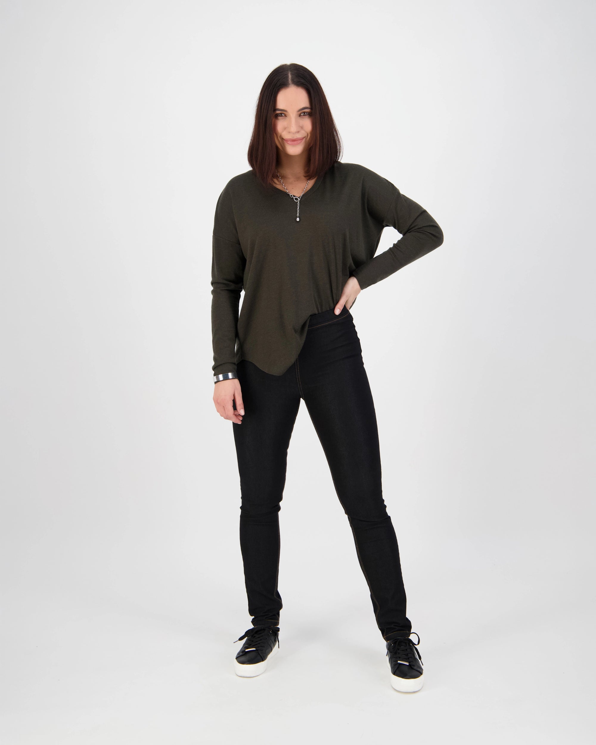A woman with straight, shoulder-length brown hair stands confidently against a white background, wearing a dark green long-sleeve top, black sneakers with white soles, and Vassalli Origin Pull On - Black Denim (Contrast Stitch) jeans. One hand is on her hip.