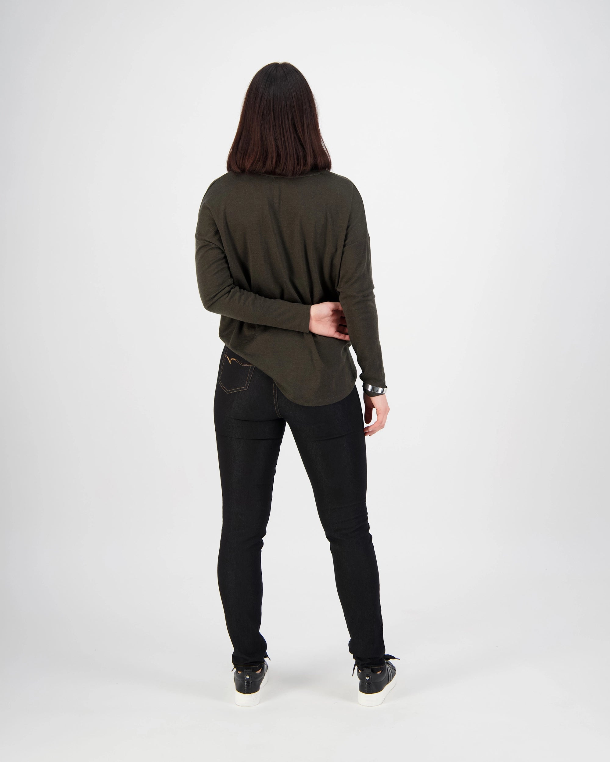 A person with shoulder-length dark hair stands facing away, wearing a dark top and Vassalli Origin Pull On - Black Denim (Contrast Stitch) jeans. Their right arm is bent behind their back, holding their left elbow. The background is plain white.