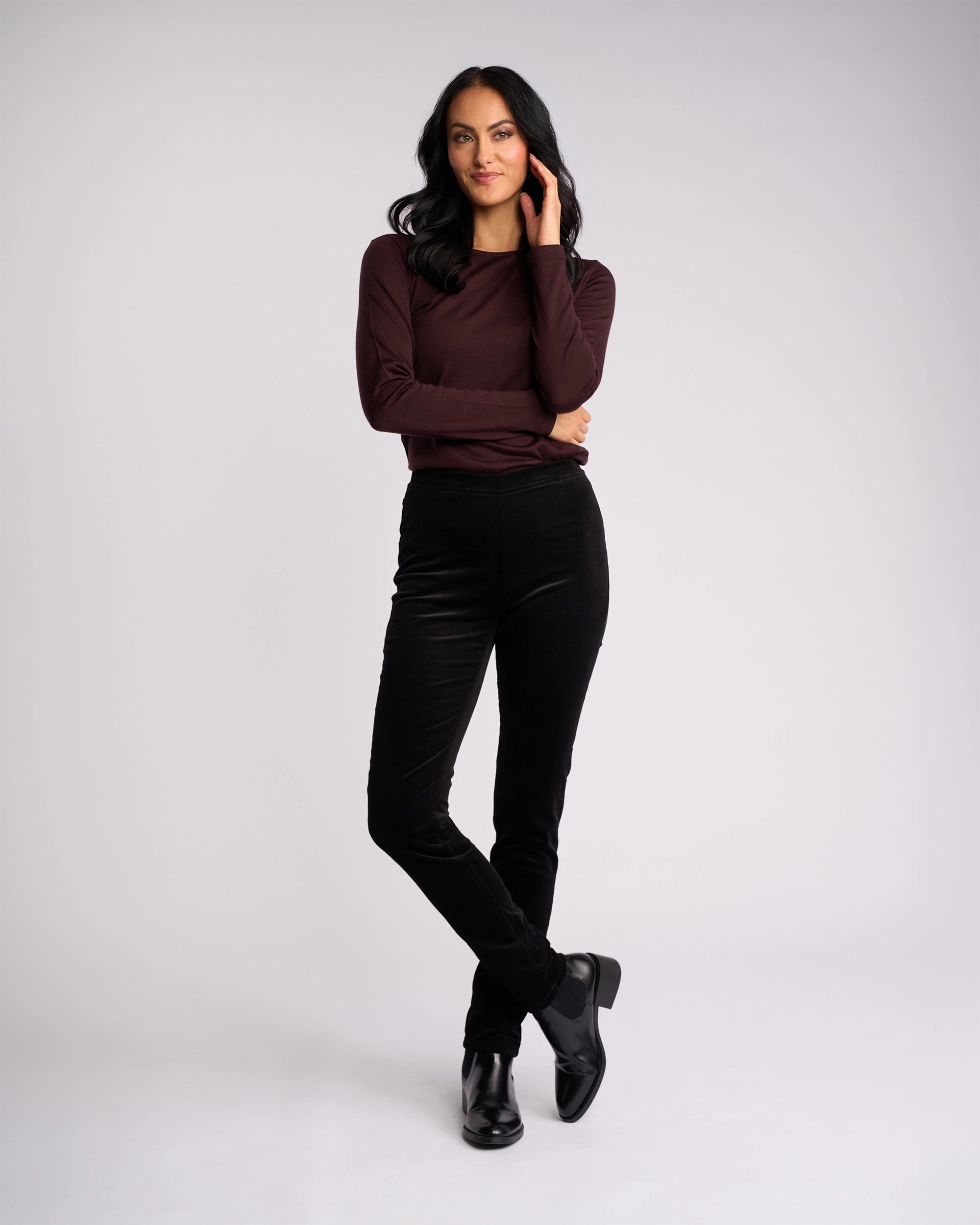 A woman with long dark hair, wearing a maroon long-sleeve top, Vassalli Origin Cord Pull On - Black skinny-leg pants, and black boots stands confidently against a plain light background with one hand in her hair and one leg crossed.