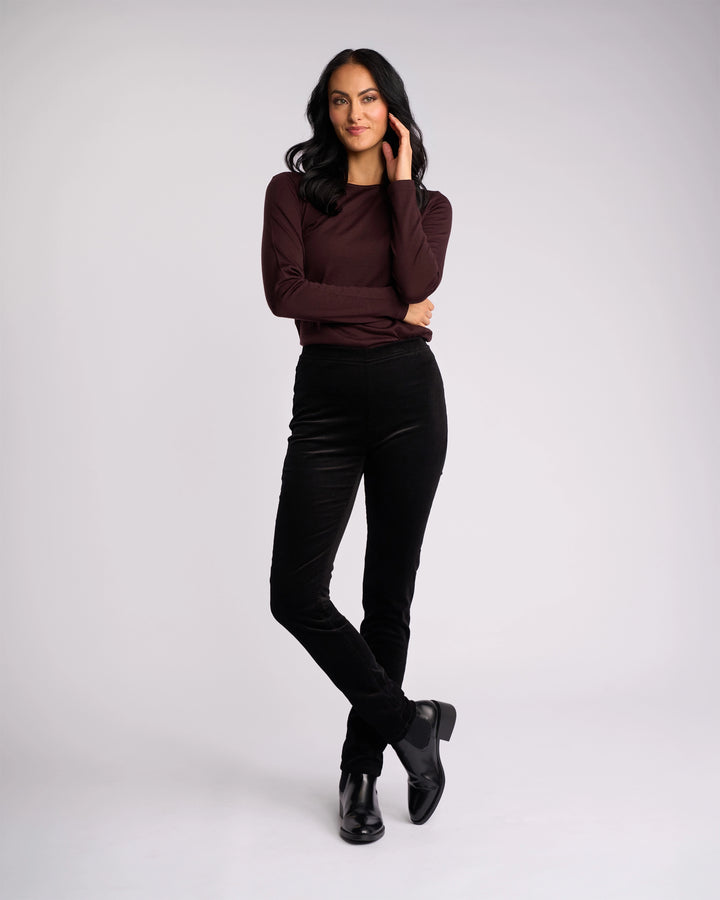 A woman with long dark hair, wearing a maroon long-sleeve top, Vassalli Origin Cord Pull On - Black skinny-leg pants, and black boots stands confidently against a plain light background with one hand in her hair and one leg crossed.