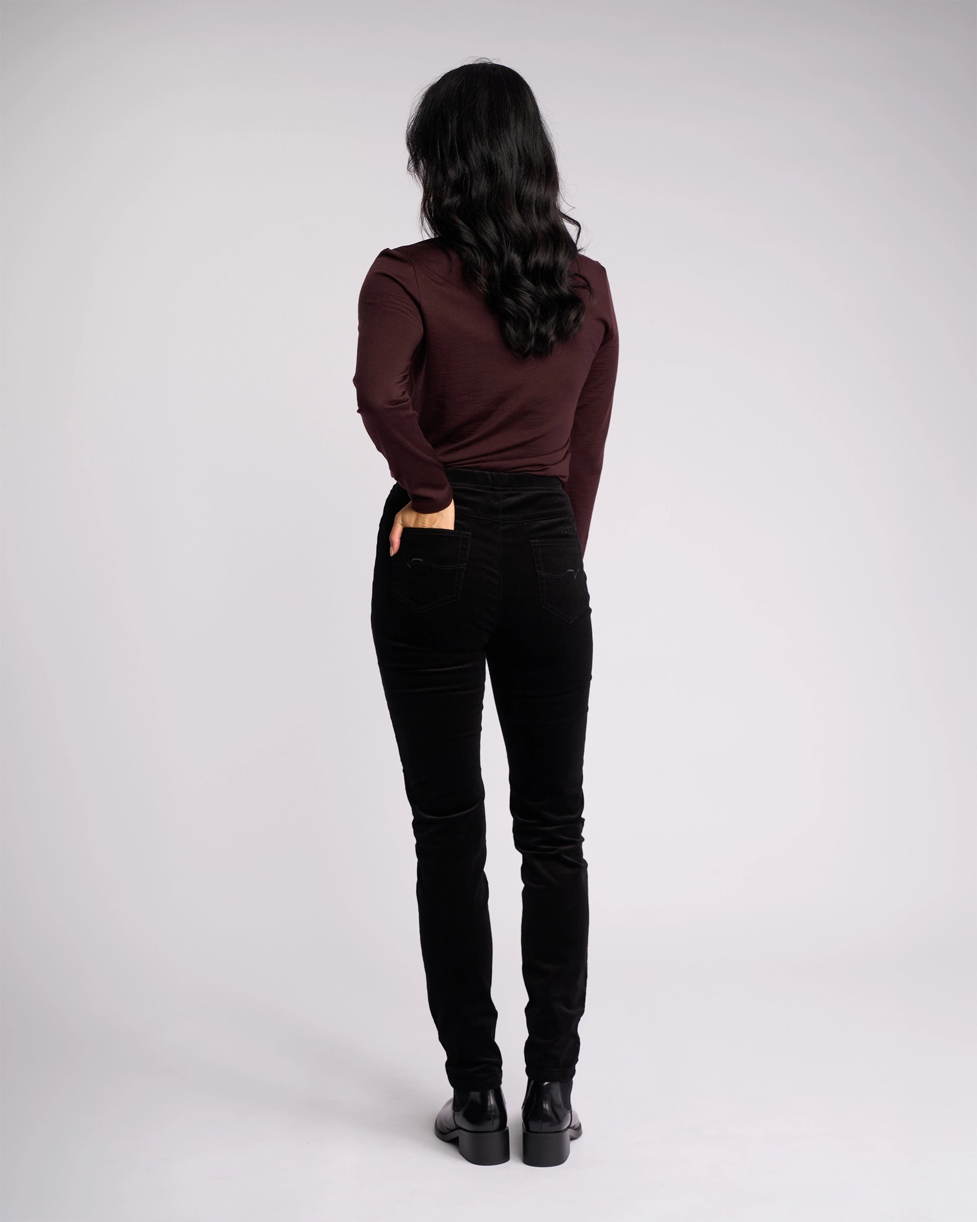 A person with long dark hair, wearing a maroon long-sleeve top and Vassalli Origin Cord Pull On - Black pants, stands facing away with one hand in their back pocket against a plain light background.