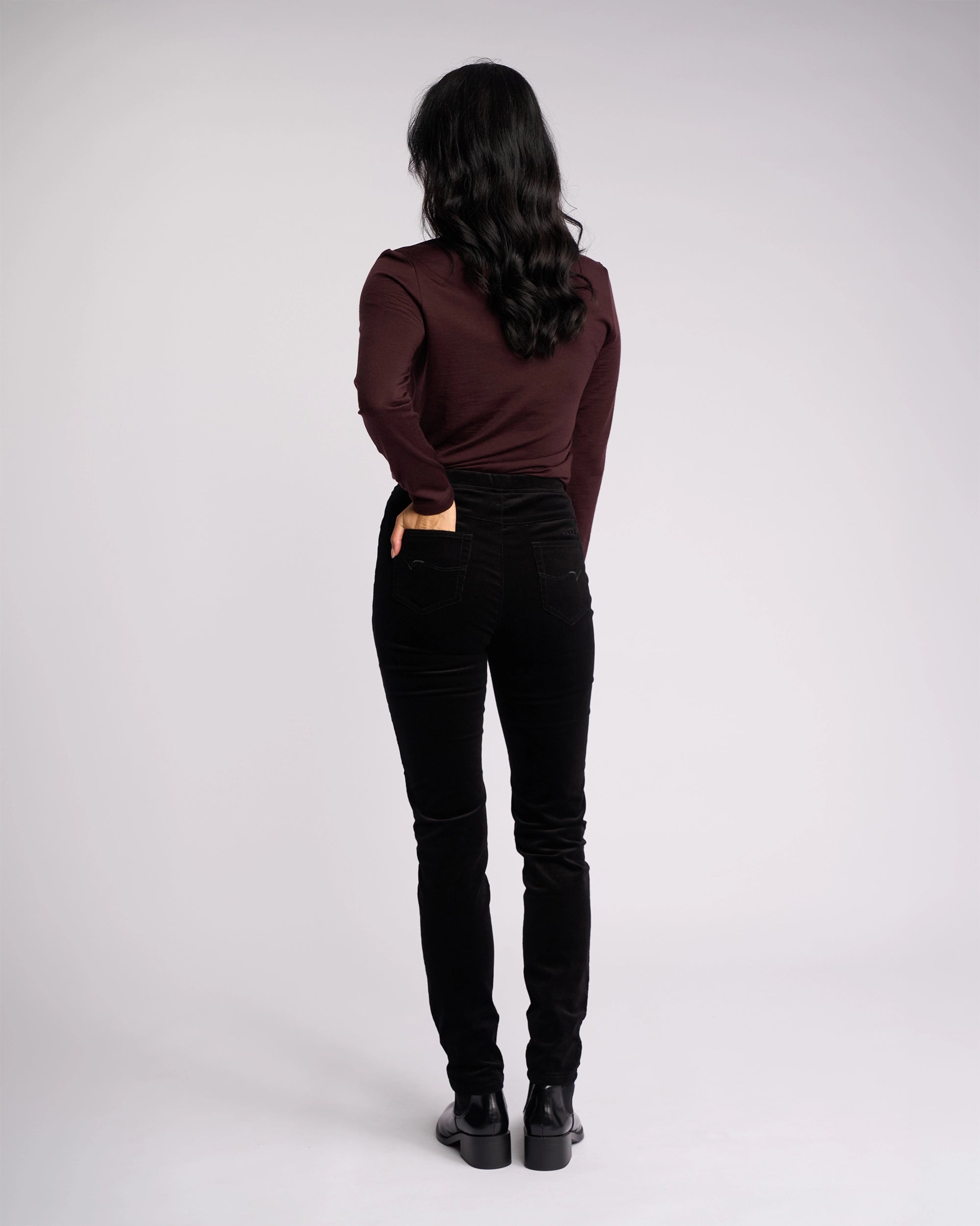 A person with long dark hair, wearing a maroon long-sleeve top and Vassalli Origin Cord Pull On - Black pants, stands facing away with one hand in their back pocket against a plain light background.