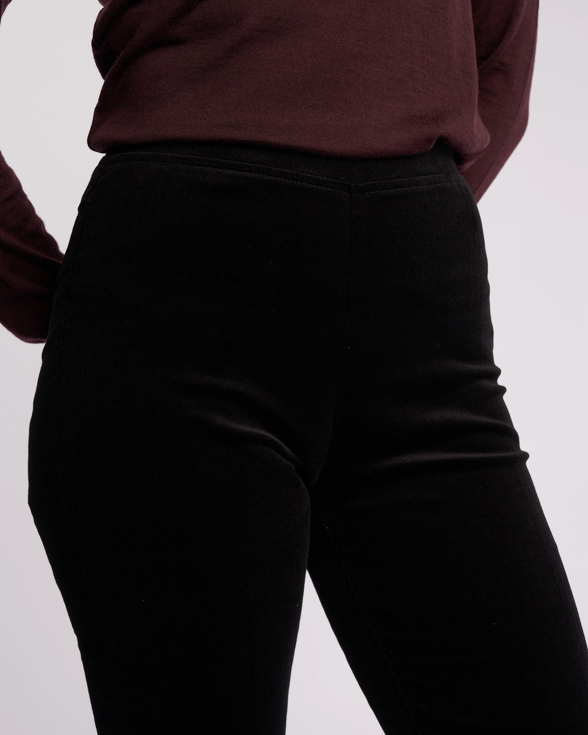 A person wearing the Vassalli Origin Cord Pull On - Black, a pair of high-waisted, skinny-leg stretch cord pants, is shown from shoulders to mid-thigh against a plain light background.