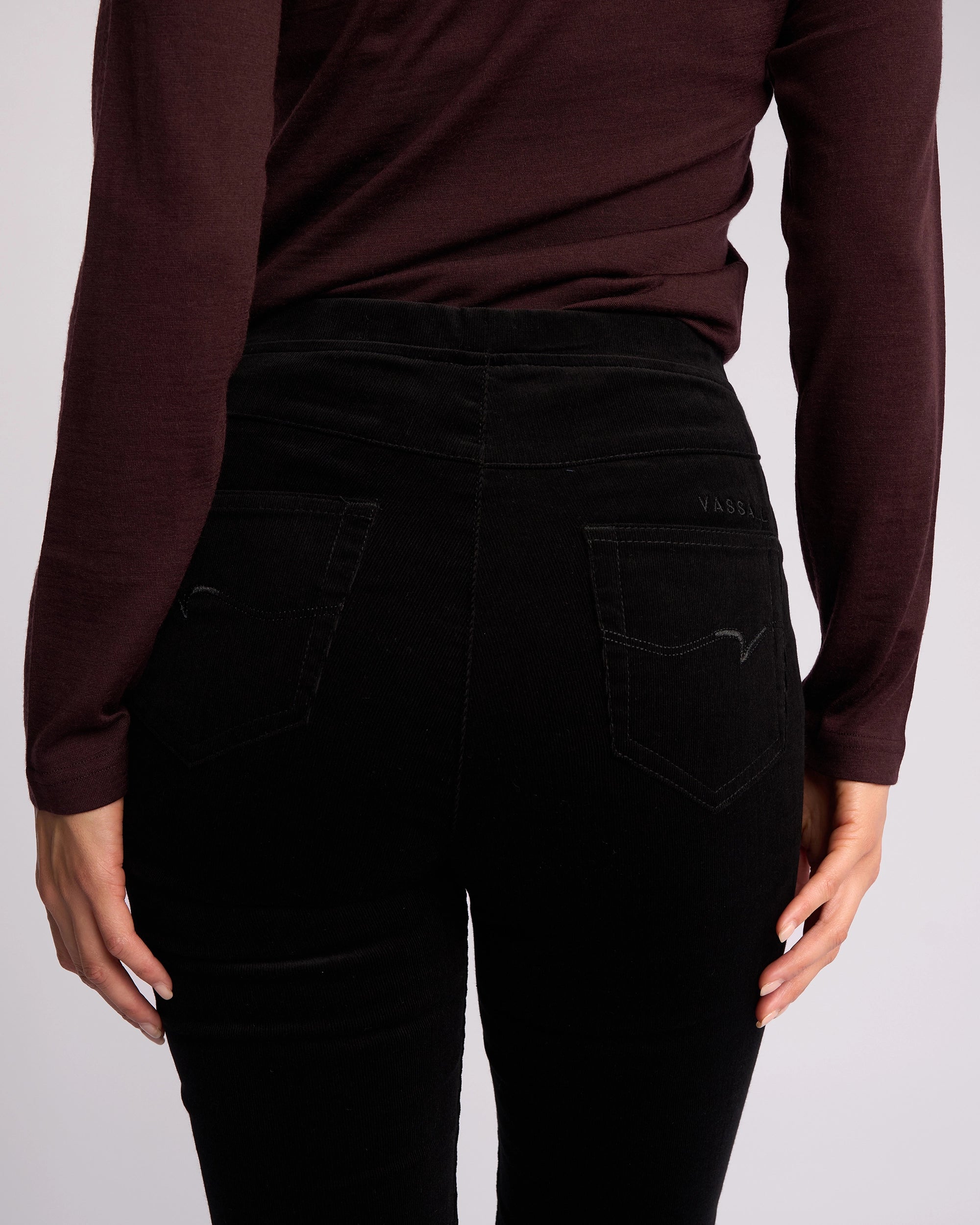A person is shown from the back wearing a long-sleeve, dark burgundy top paired with Vassalli Origin Cord Pull On skinny-leg pants in black, featuring a high waist and two back pockets, hands resting by their sides.