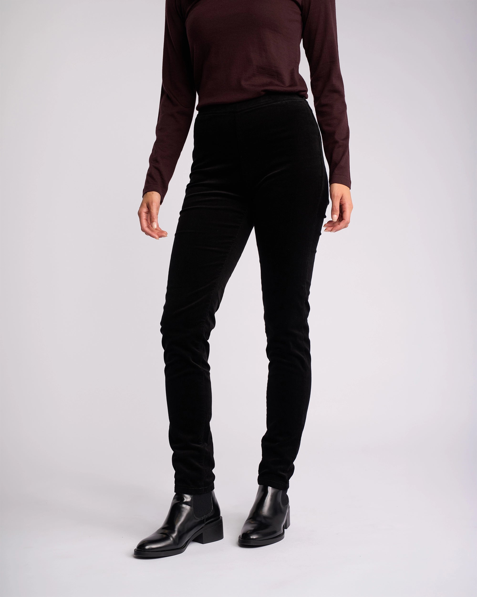 A person models Vassalli’s Origin Cord Pull On - Black pants, paired with a long-sleeve dark top and black ankle boots against a plain light background. The person’s face is not visible.