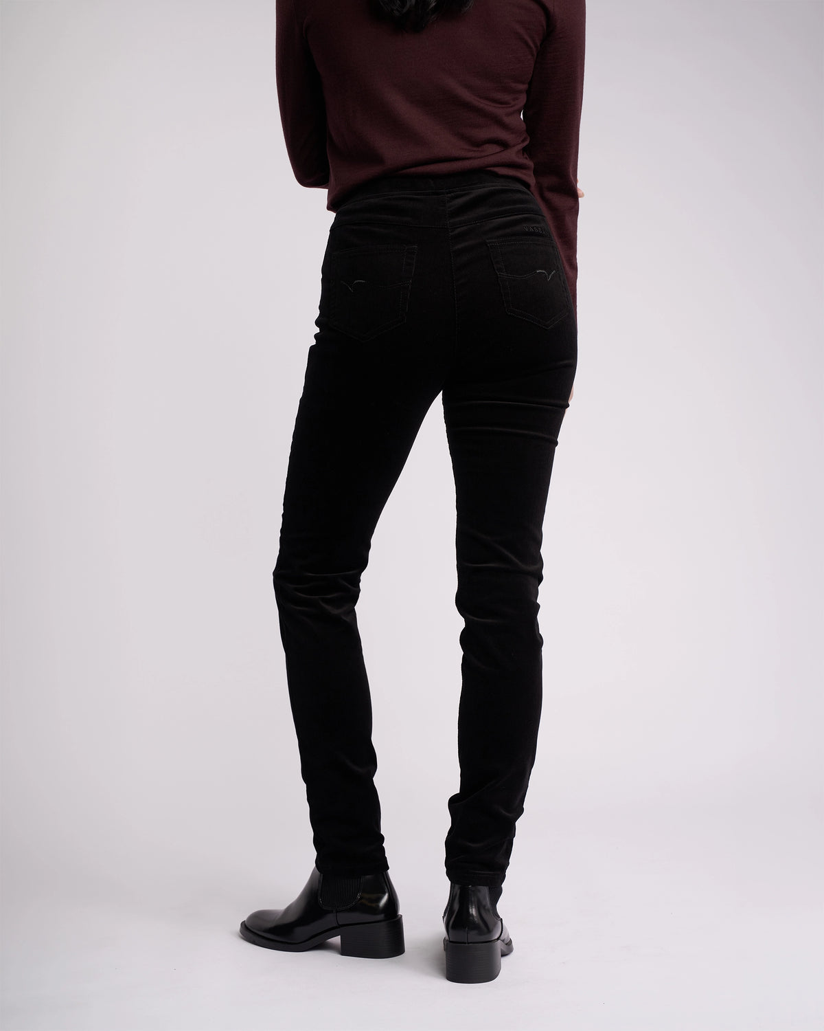 A person is shown from the back, shoulders down, wearing a maroon long-sleeve top and Vassalli Origin Cord Pull On - Black skinny-leg pants with ankle boots, set against a plain white background.