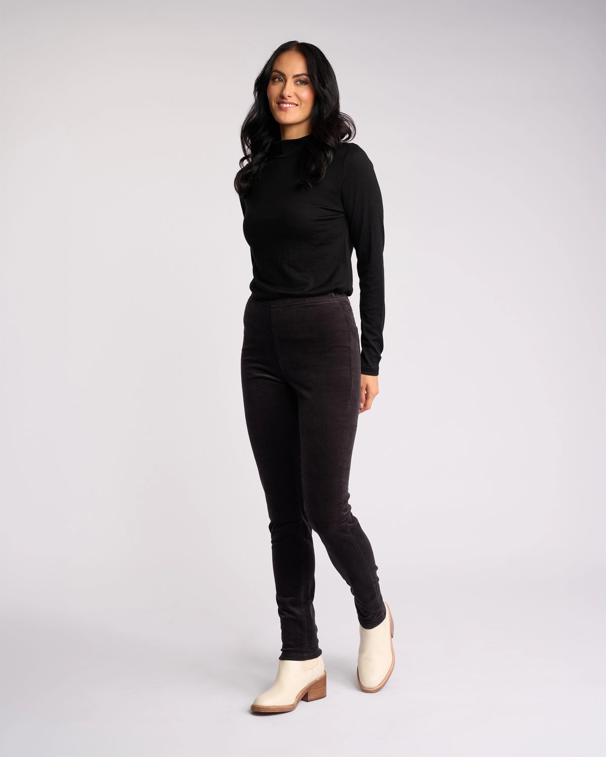 A woman with long dark hair stands against a plain light background, smiling with one foot slightly forward. She wears a black long-sleeve top, Vassalli Origin Cord Pull On pants in charcoal, and white heeled ankle boots.