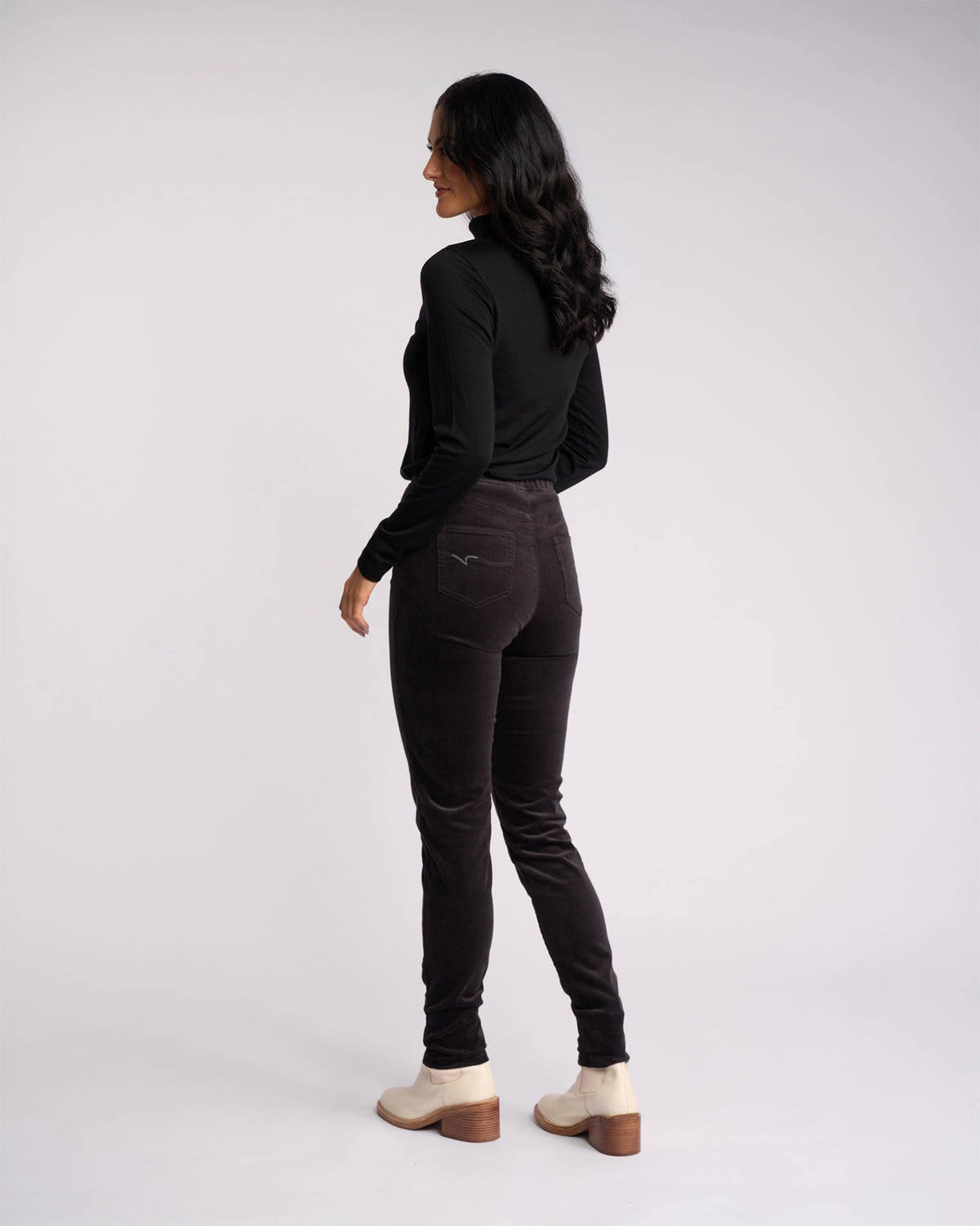 A woman with long dark hair wears a black long-sleeve top, light-colored shoes, and Vassalli’s Origin Cord Pull On - Charcoal skinny-leg pants, standing sideways against a plain light background.