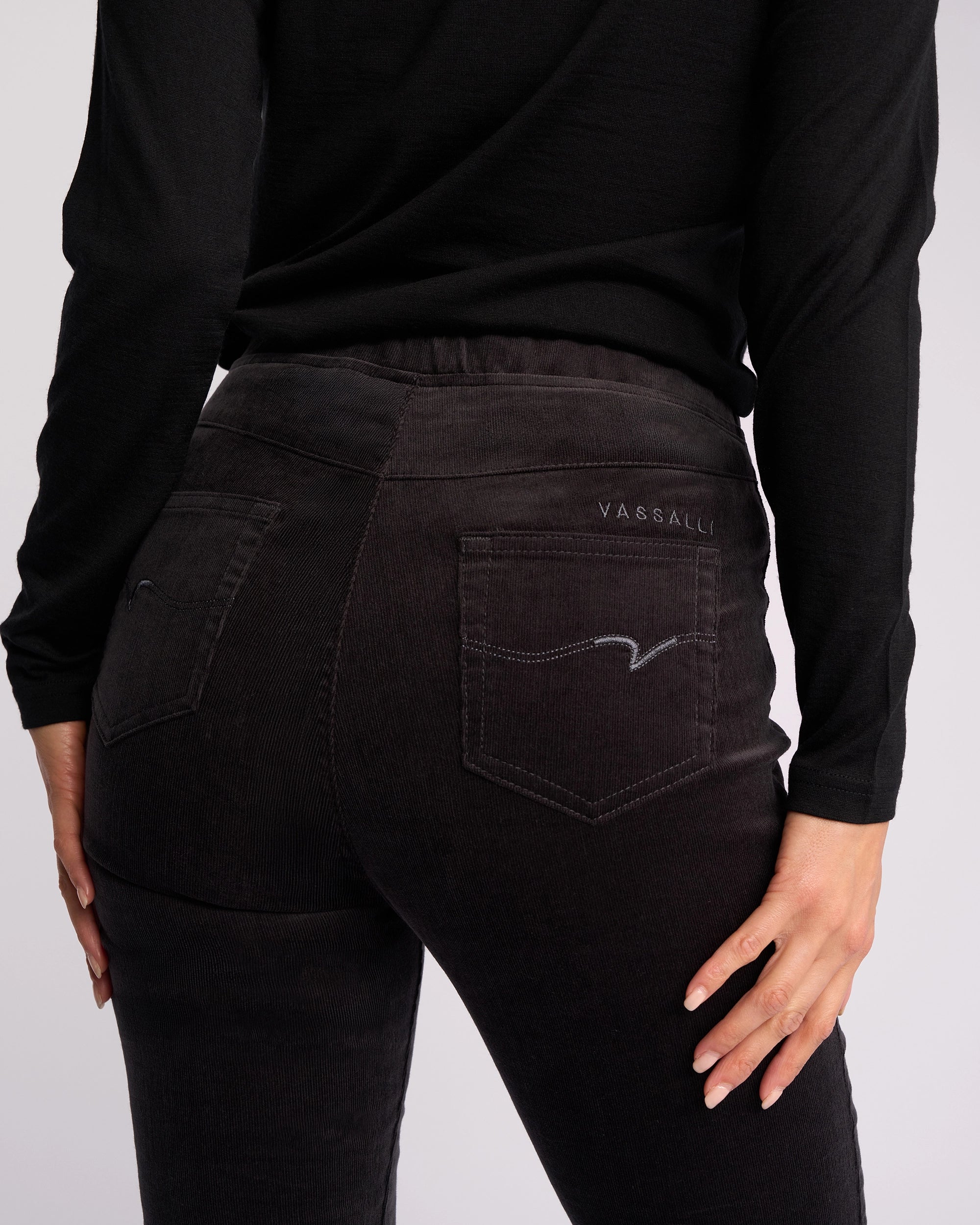 A person wearing Vassalli's Origin Cord Pull On pants in charcoal and a black long-sleeve top, shown from the back. The pants have two back pockets, one with “Vassalli” stitched above it. Their right hand rests on their hip.