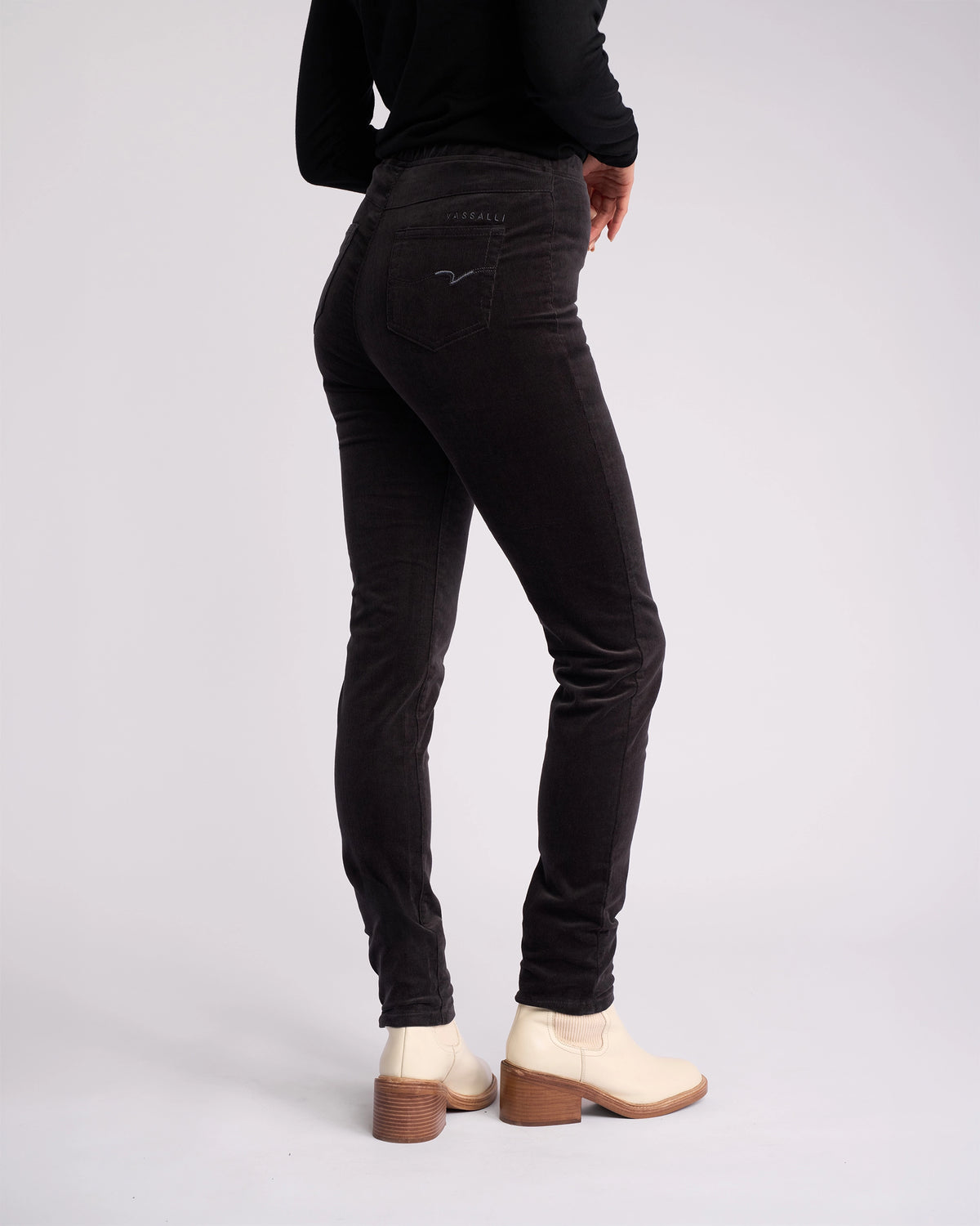 Person stands against a plain background, wearing Vassalli Origin Cord Pull On pants in Charcoal, a black long-sleeve top, and cream ankle boots with wooden heels; facing away with head out of frame.