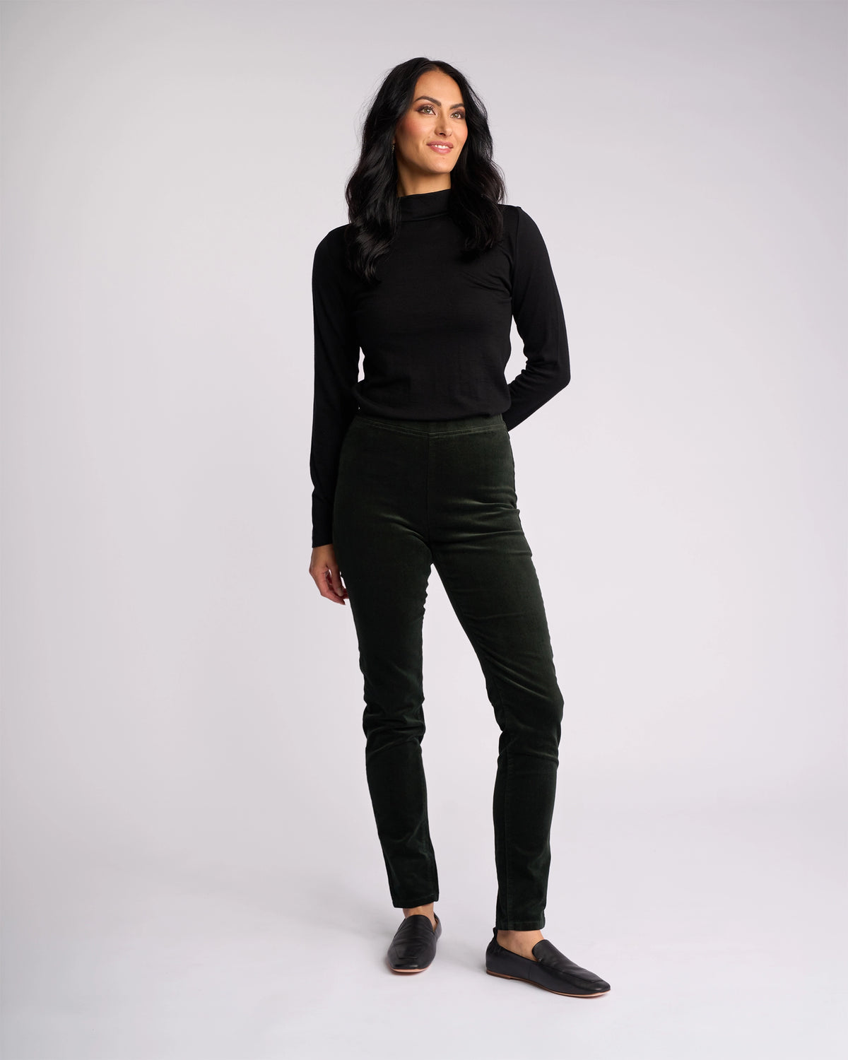 A woman stands against a plain light background, wearing the Vassalli Origin Cord Pull On - Forest pants with a long-sleeve black top and black slip-on shoes. Her left hand is by her side while her right rests on her thigh.