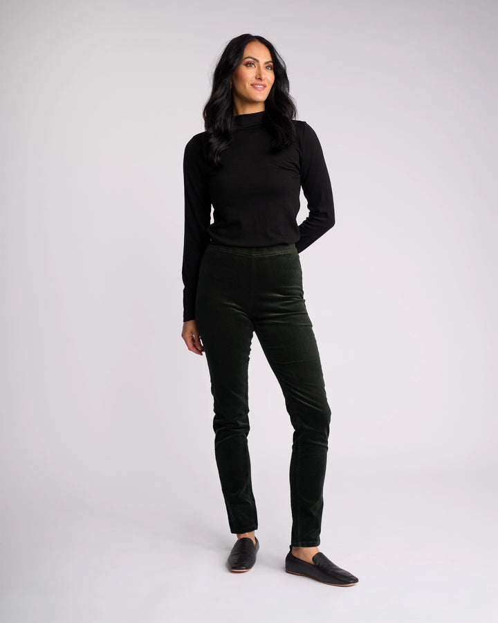 A woman stands against a plain light background, wearing the Vassalli Origin Cord Pull On - Forest pants with a long-sleeve black top and black slip-on shoes. Her left hand is by her side while her right rests on her thigh.