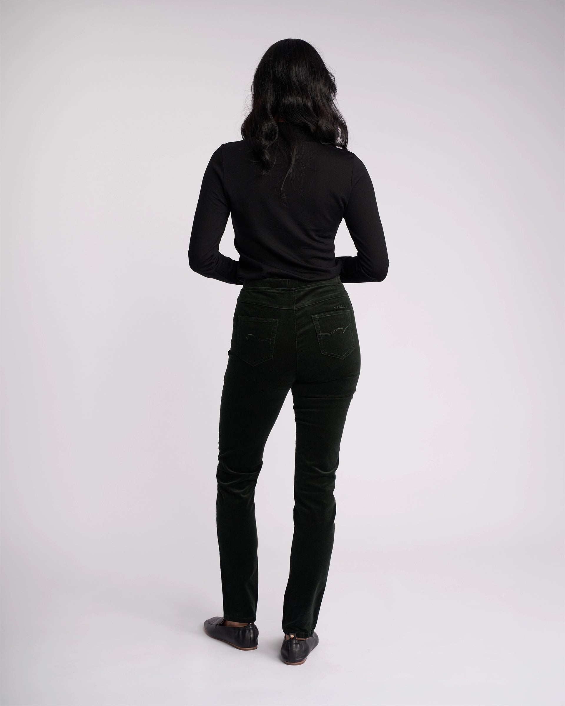 A person with long dark hair stands facing away, wearing a black long-sleeve top, black shoes, and Vassalli Origin Cord Pull On - Forest pants, against a plain light background.