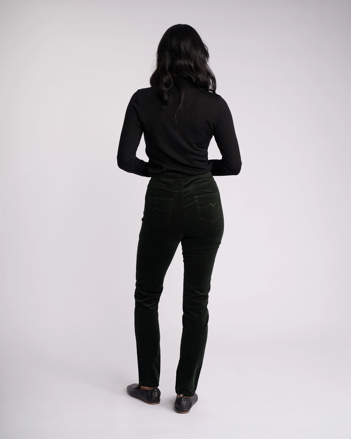 A person with long dark hair stands facing away, wearing a black long-sleeve top, black shoes, and Vassalli Origin Cord Pull On - Forest pants, against a plain light background.