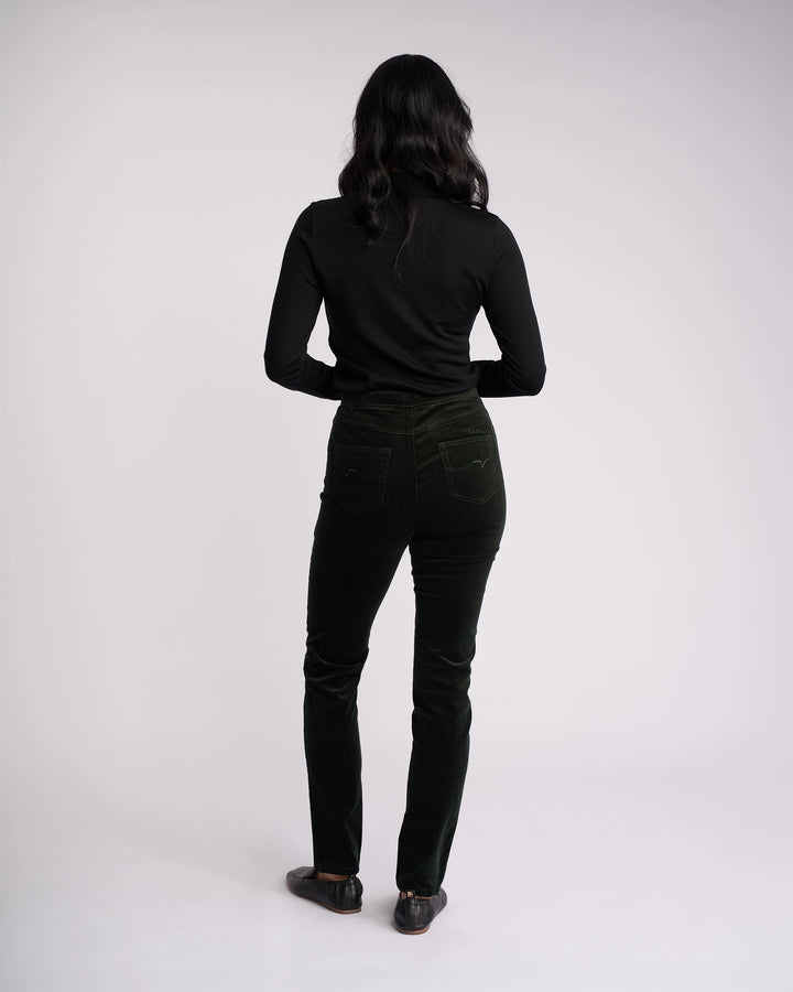 A person with long dark hair stands facing away, wearing a black long-sleeve top, black shoes, and Vassalli Origin Cord Pull On - Forest pants, against a plain light background.