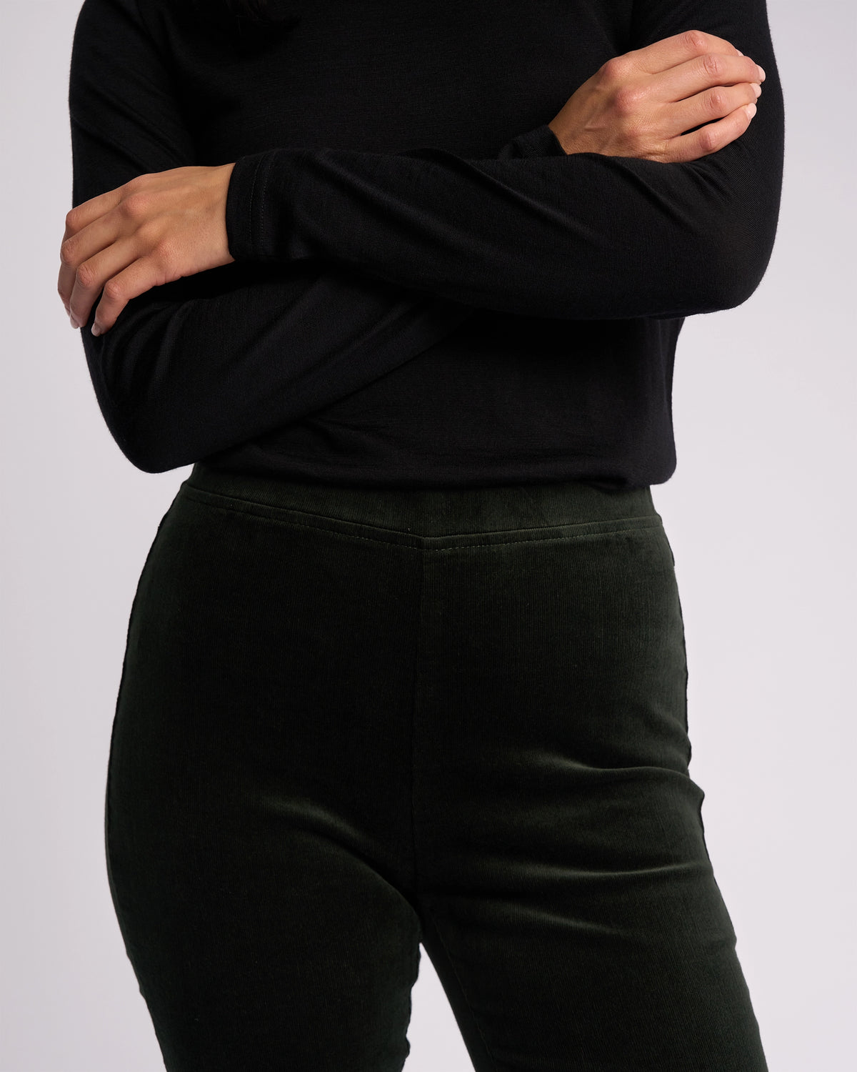A person models the Vassalli Origin Cord Pull On - Forest, a dark green skinny-leg pant with a pull-on design, paired with a long-sleeved black top, shown from shoulders to thighs against a plain light background.