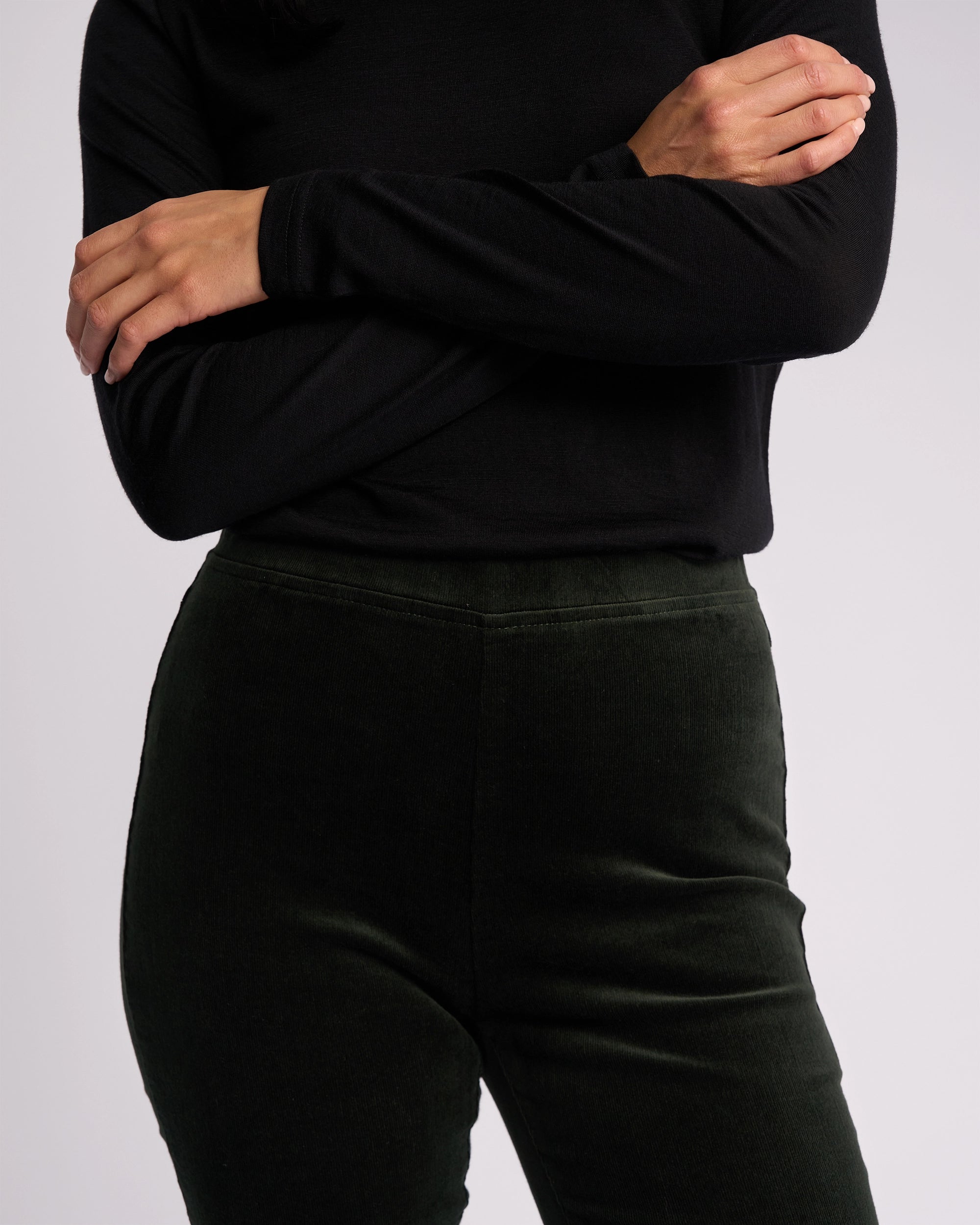 A person models the Vassalli Origin Cord Pull On - Forest, a dark green skinny-leg pant with a pull-on design, paired with a long-sleeved black top, shown from shoulders to thighs against a plain light background.