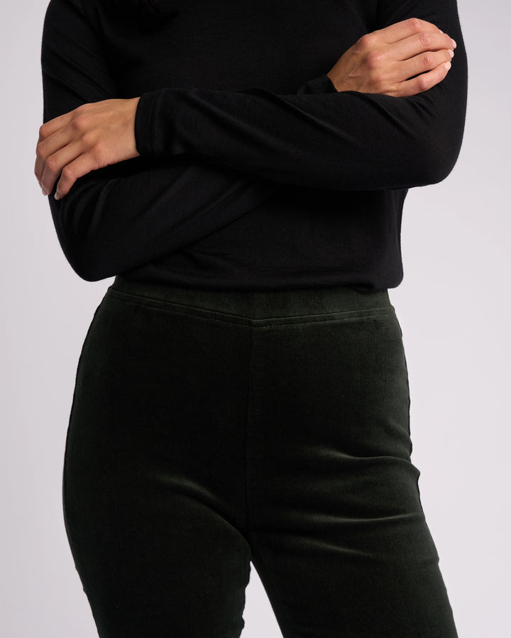 A person models the Vassalli Origin Cord Pull On - Forest, a dark green skinny-leg pant with a pull-on design, paired with a long-sleeved black top, shown from shoulders to thighs against a plain light background.