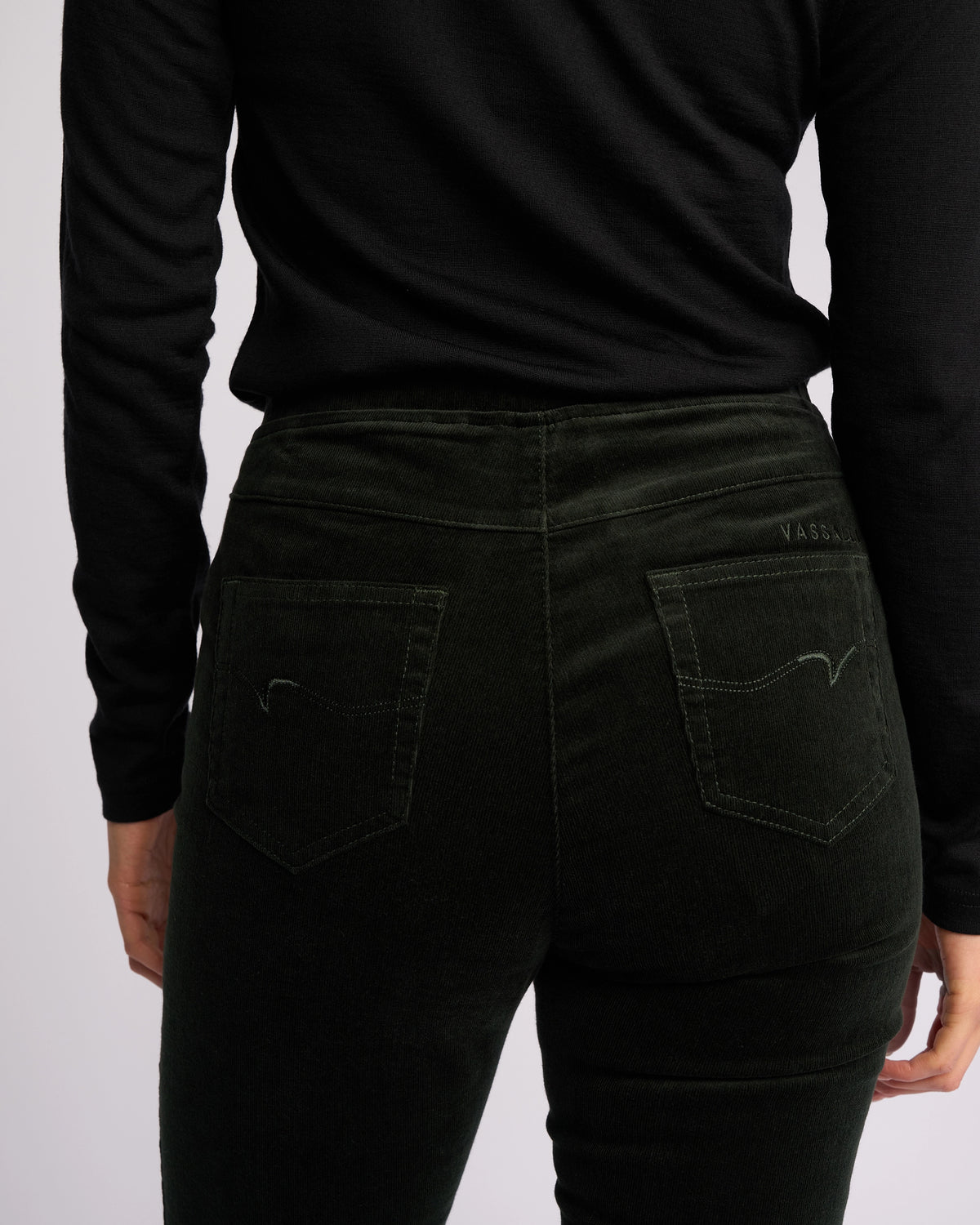 A person is shown from the back wearing Vassalli's Origin Cord Pull On - Forest pants, highlighting the back pockets and curved seam details, paired with a fitted black long-sleeve top.