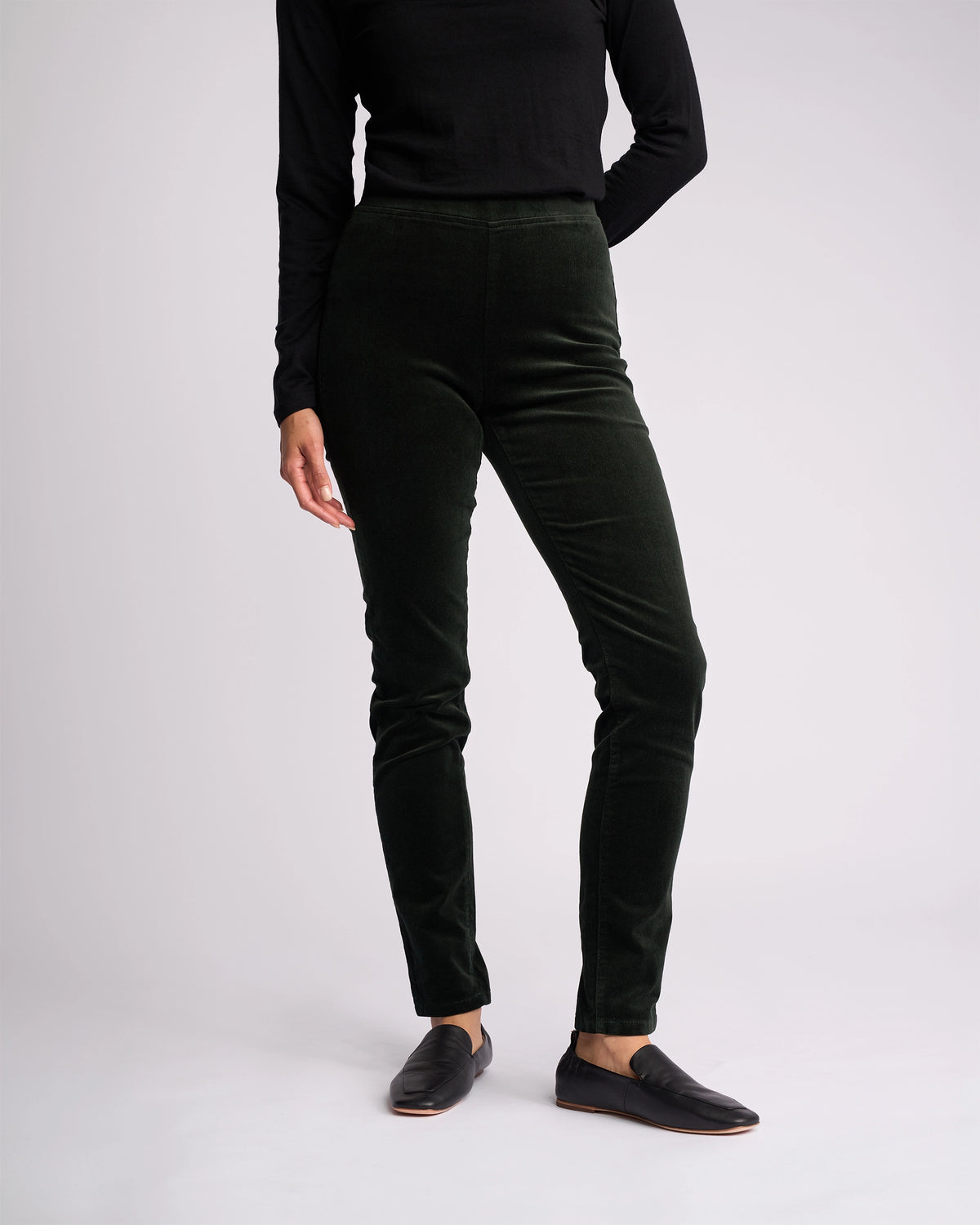 A person stands against a plain light background wearing a fitted black long-sleeve top, Vassalli Origin Cord Pull On - Forest pants, and black slip-on shoes. Their face is not visible.