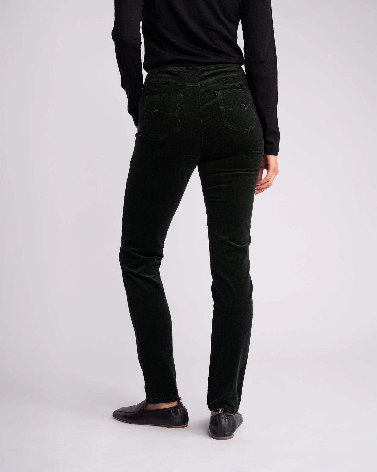 A person stands facing away, showcasing the back of the Vassalli Origin Cord Pull On - Forest pants with a black long-sleeve top and black flats against a plain light background.