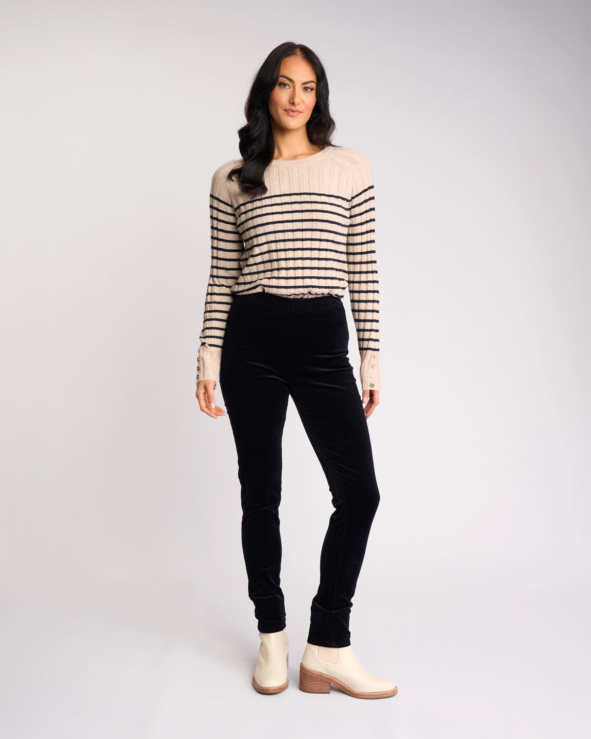 A woman with long, dark wavy hair stands facing forward against a plain light background, wearing the Vassalli Origin Cord Pull On pants in Midnight, a beige sweater with black stripes, and cream ankle boots.
