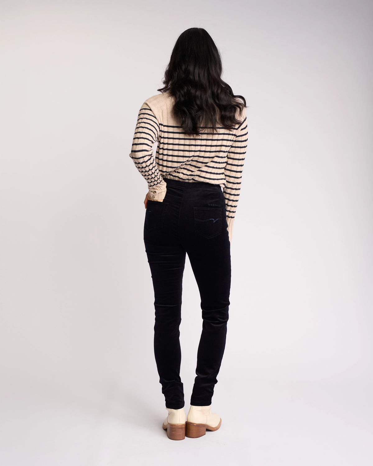 A person with long dark hair, facing away from the camera against a plain white background, wears a beige and black striped sweater and Vassalli Origin Cord Pull On - Midnight skinny-leg pants with light-colored shoes.