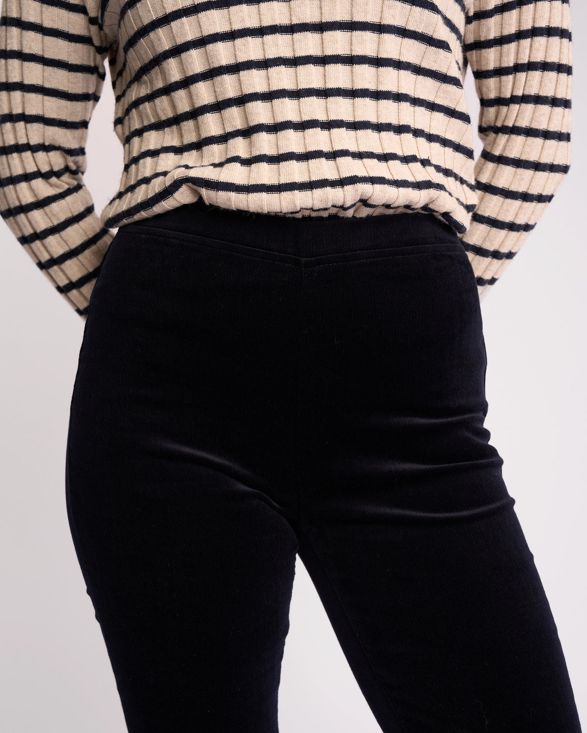 A person wears a beige and black striped long-sleeve sweater tucked into Vassalli Origin Cord Pull On - Midnight high-waisted, skinny-leg pants, shown from shoulders to mid-thigh with arms behind their back.