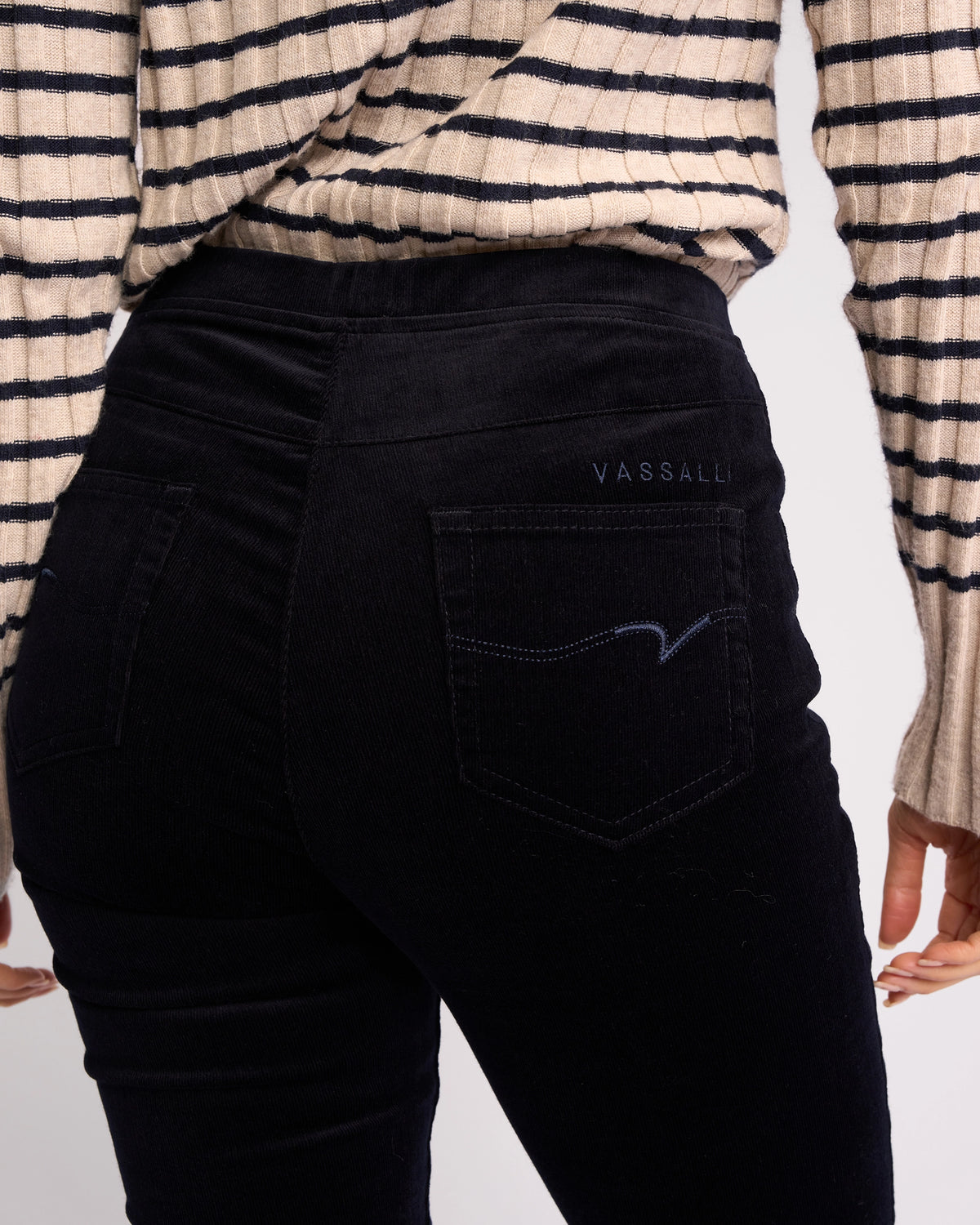 Person wearing Vassalli's Origin Cord Pull On - Midnight high-waisted skinny-leg pants, shown from the back with brand detailing on the pocket, paired with a beige and black striped sweater.