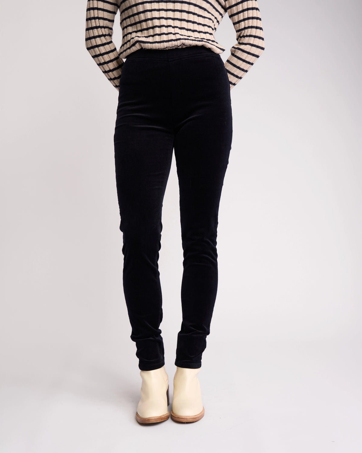 A person stands against a plain light background with hands behind their back, wearing Vassalli’s Origin Cord Pull On - Midnight high-waisted skinny-leg pants, a black and white striped long-sleeve top, and white ankle boots.