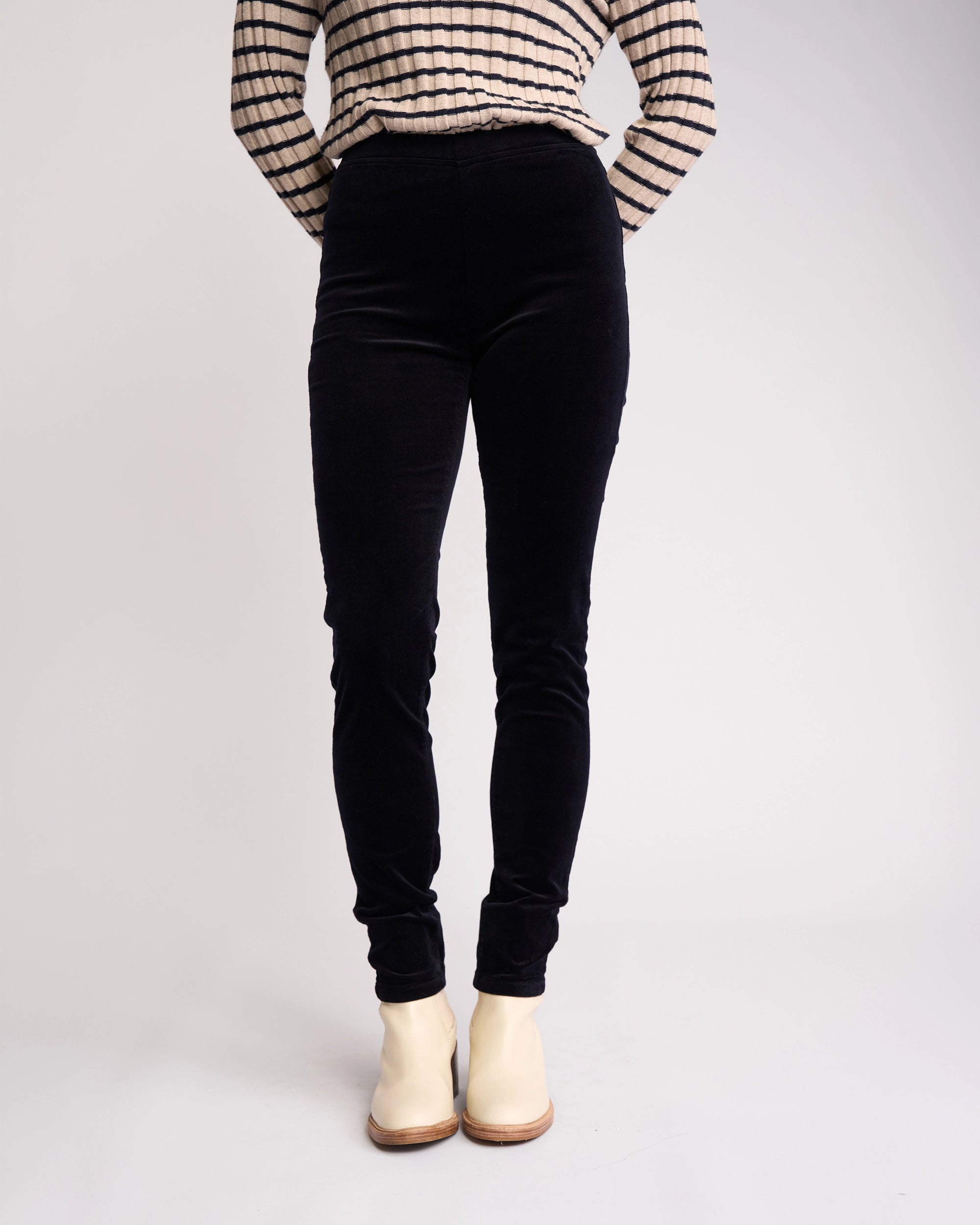 A person stands against a plain light background with hands behind their back, wearing Vassalli’s Origin Cord Pull On - Midnight high-waisted skinny-leg pants, a black and white striped long-sleeve top, and white ankle boots.