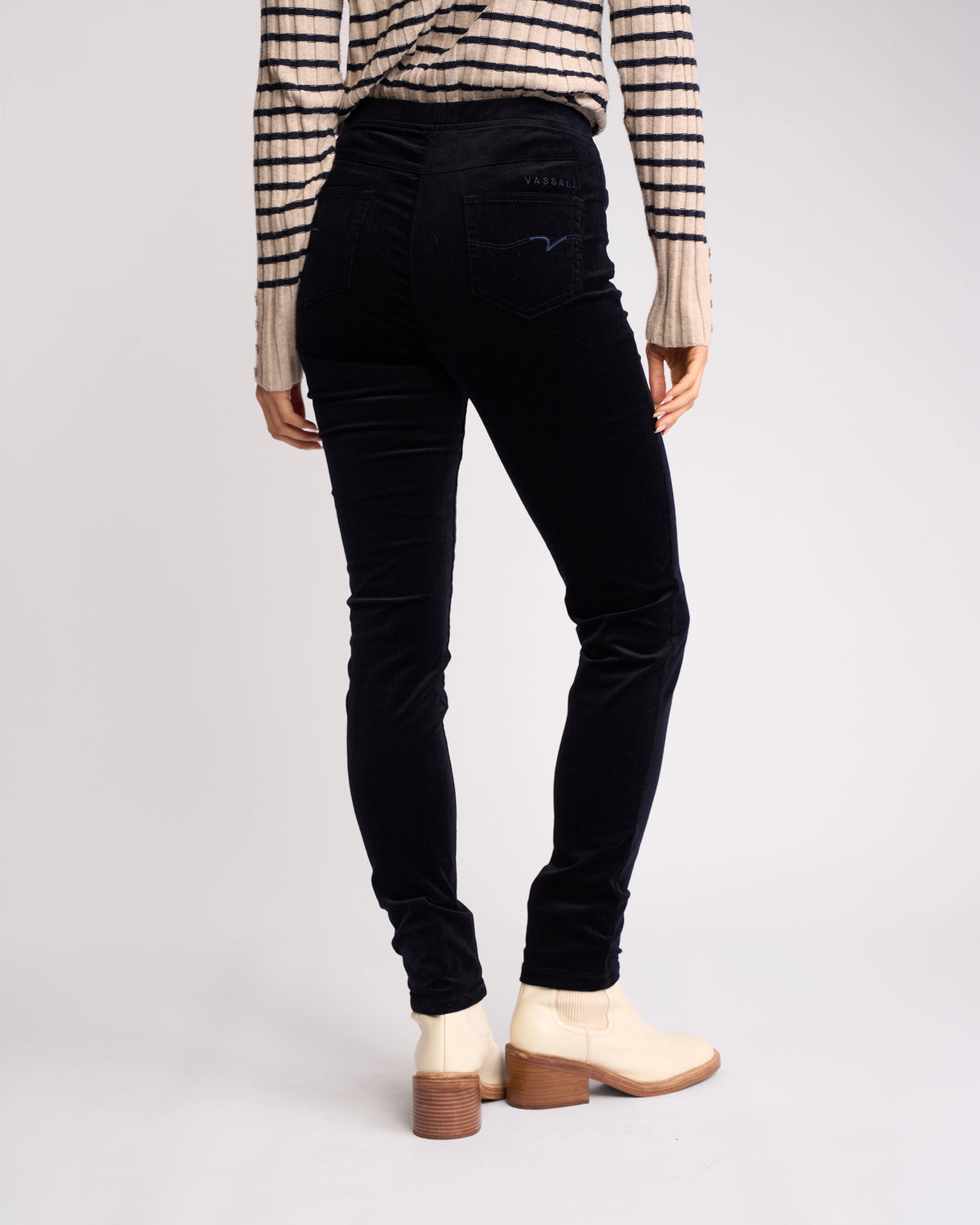 A person stands facing away against a plain light background, wearing the Vassalli Origin Cord Pull On - Midnight skinny-leg pants with a pull-on design, a beige and black striped sweater, and light-colored ankle boots.