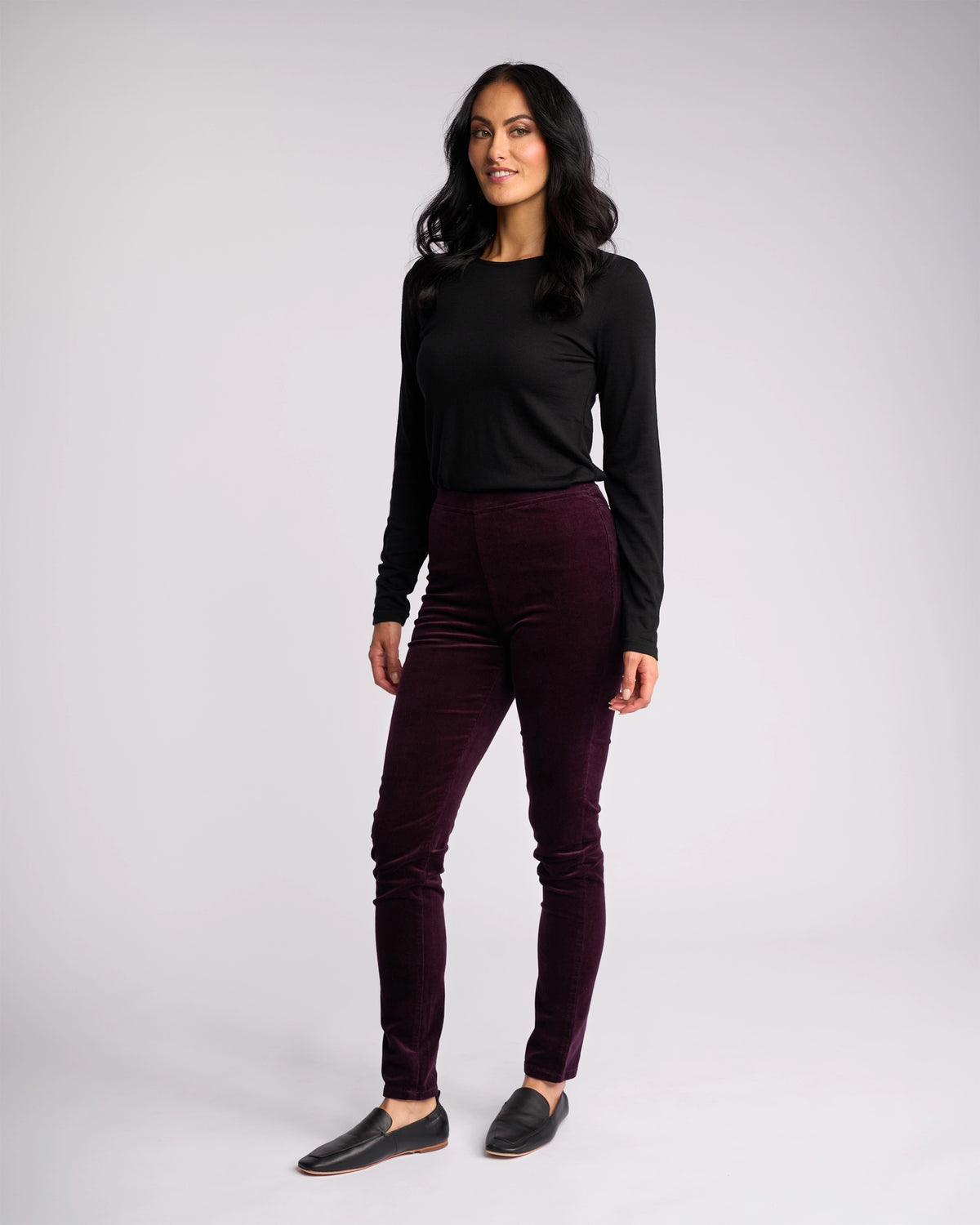 A woman with long dark hair poses against a plain background, wearing Vassalli’s Origin Cord Pull On pants in Mulberry with a black long-sleeve top and black slip-on shoes, smiling slightly as she stands with one leg forward.